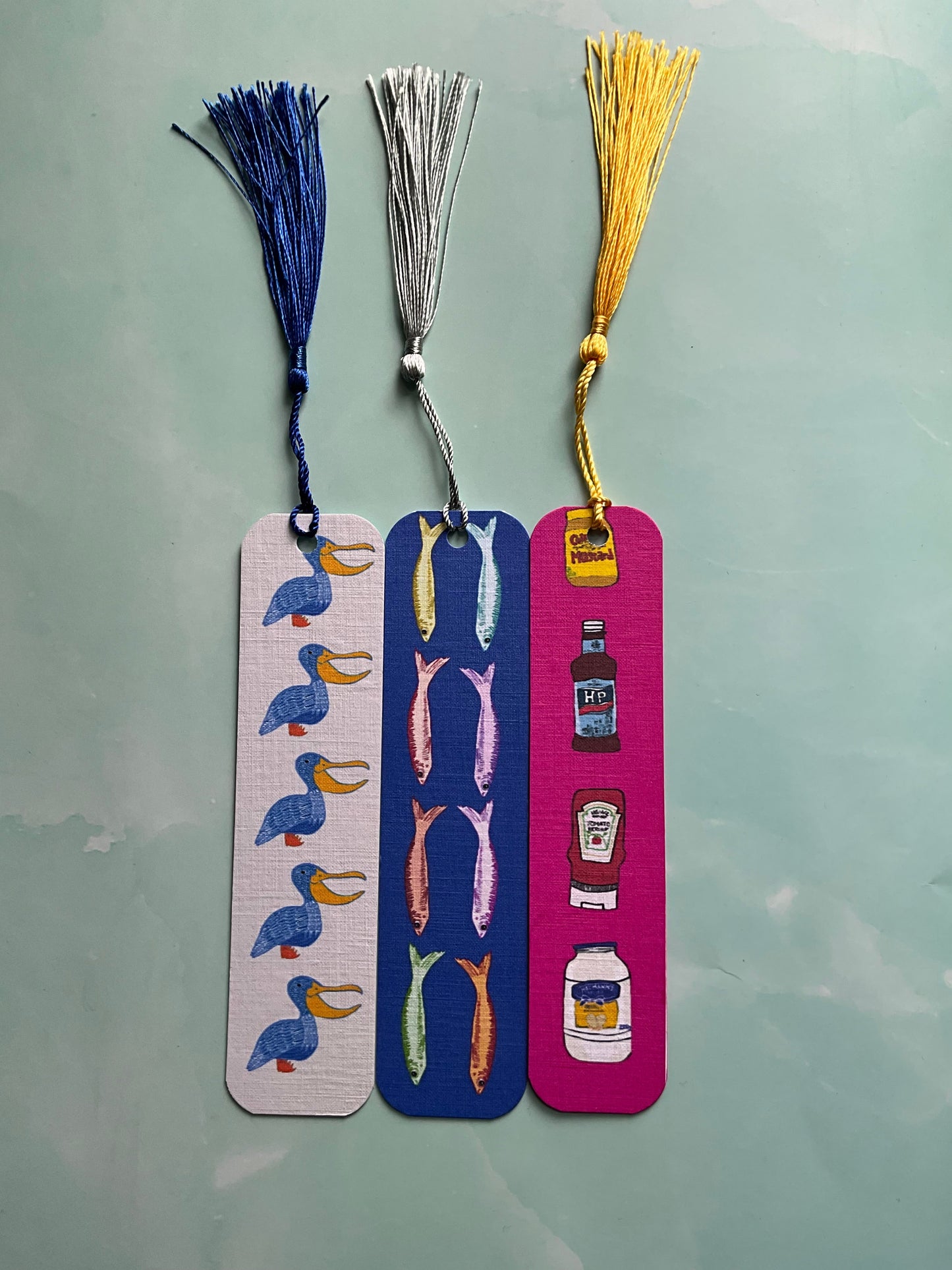 Three colorful bookmarks one featuring a bird print, one a fish print, and one a condiments print, all with tassels resting on a light blue background