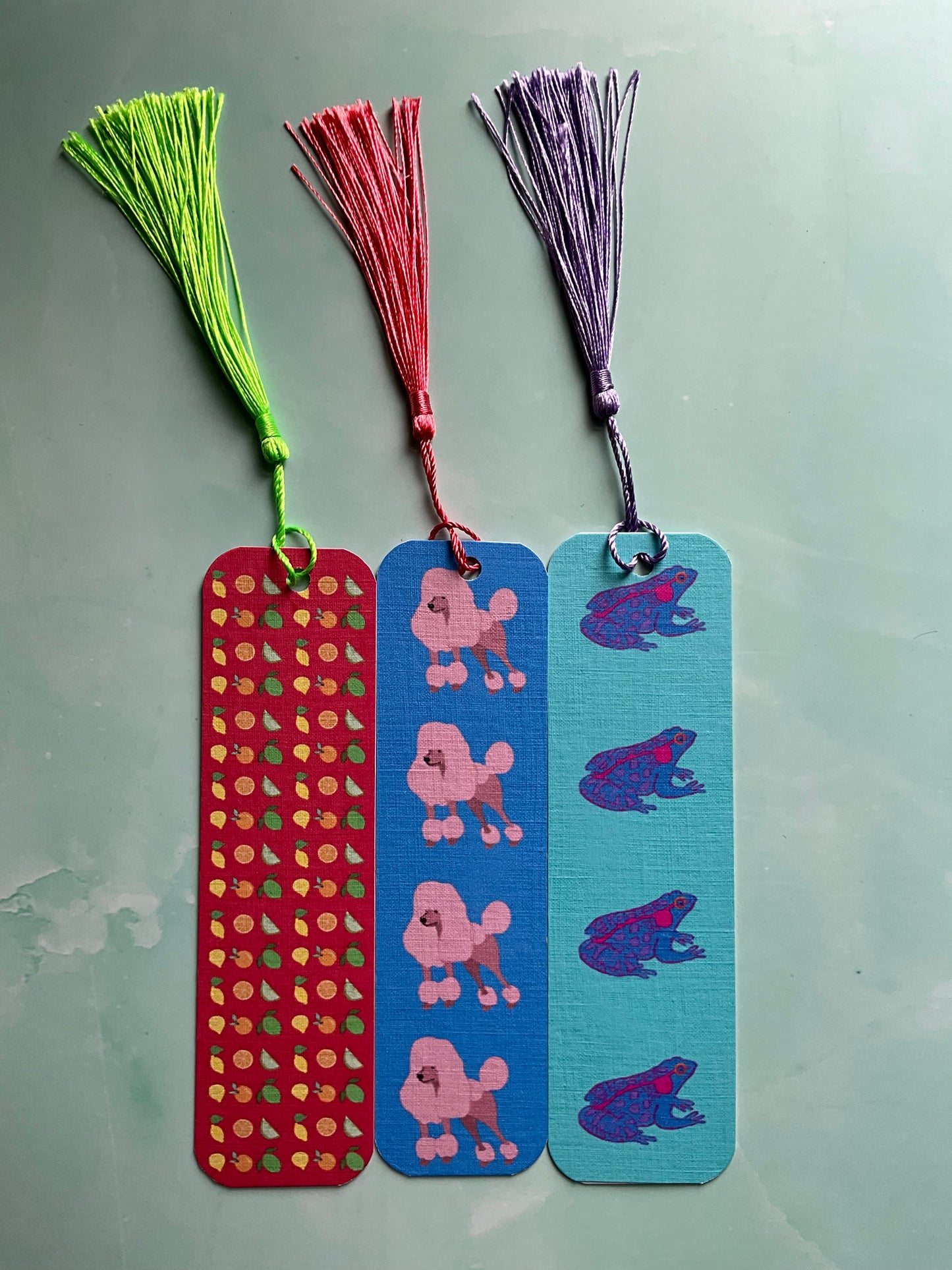 Three colourful bookmarks with tassels. One with an orange, lemon and lime print, one with a poodle print and one with a frog print.