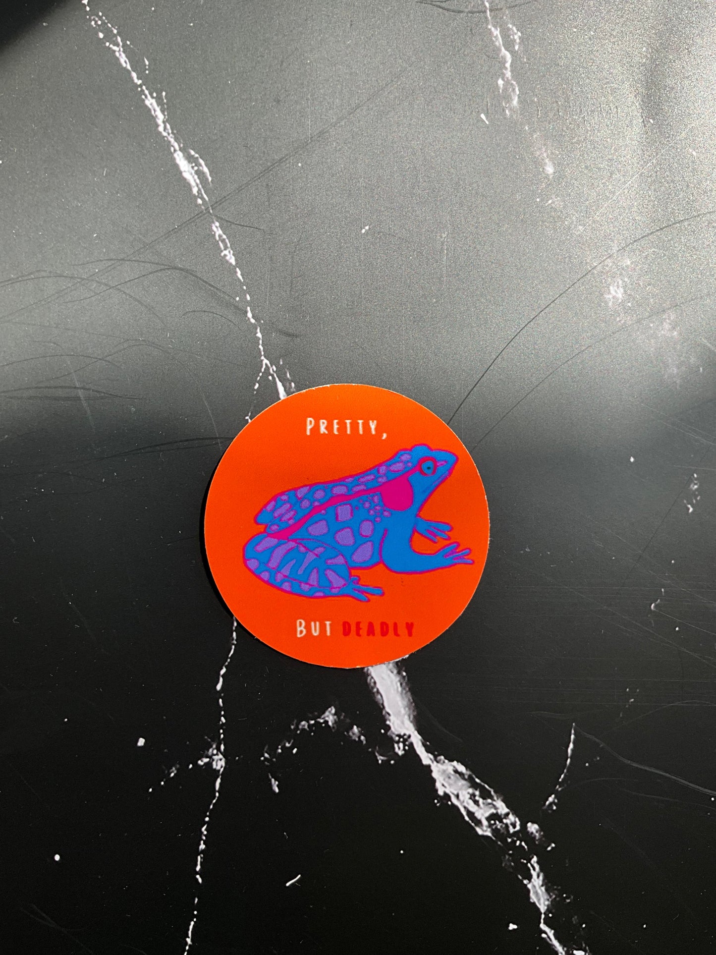 Round orange sticker with blue and pink graphic frog on a black marbled background