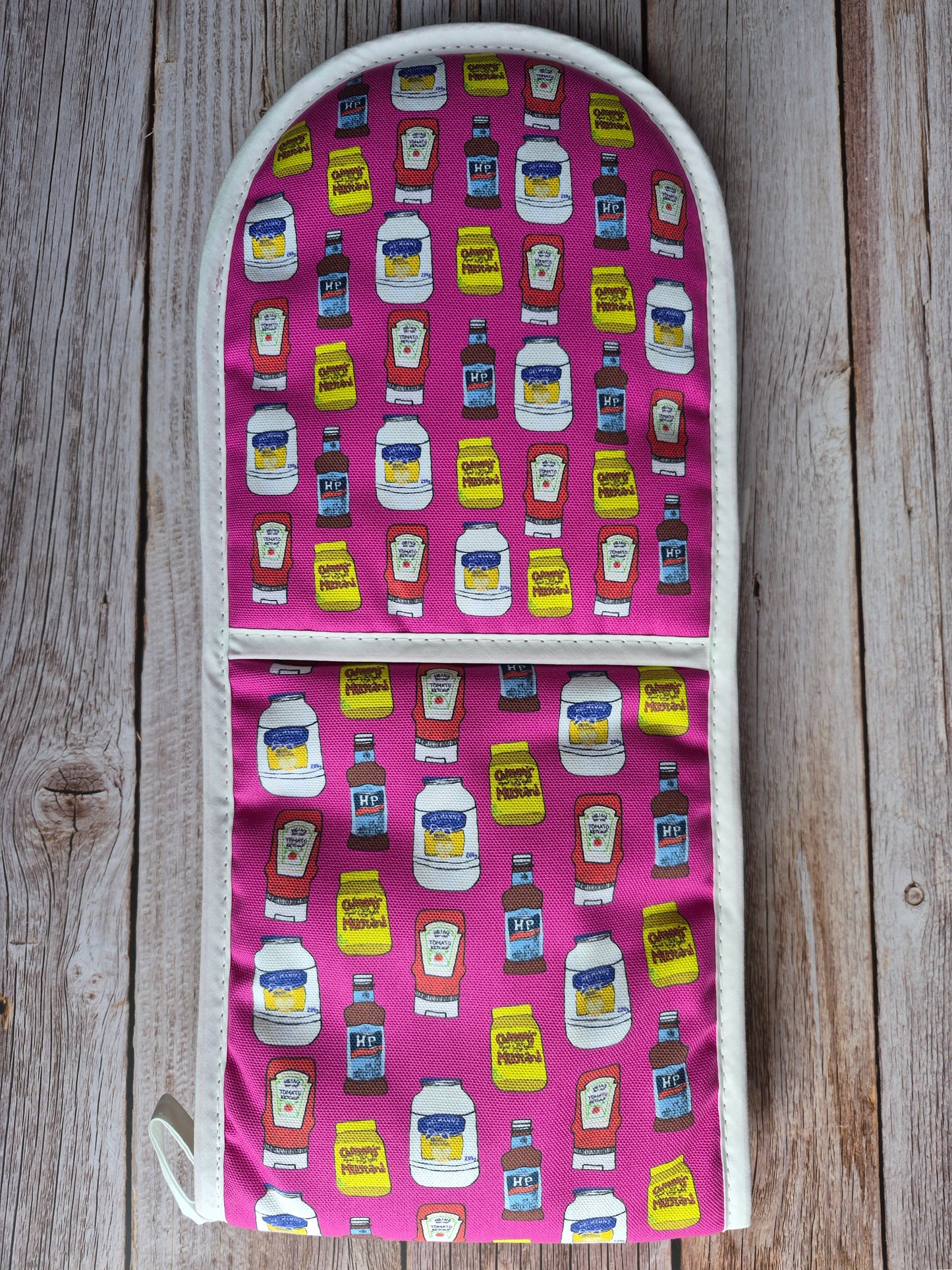 Pink oven gloves with a sauces print design (ketchup, mustard, mayo and brown sauce) on a wooden surface.