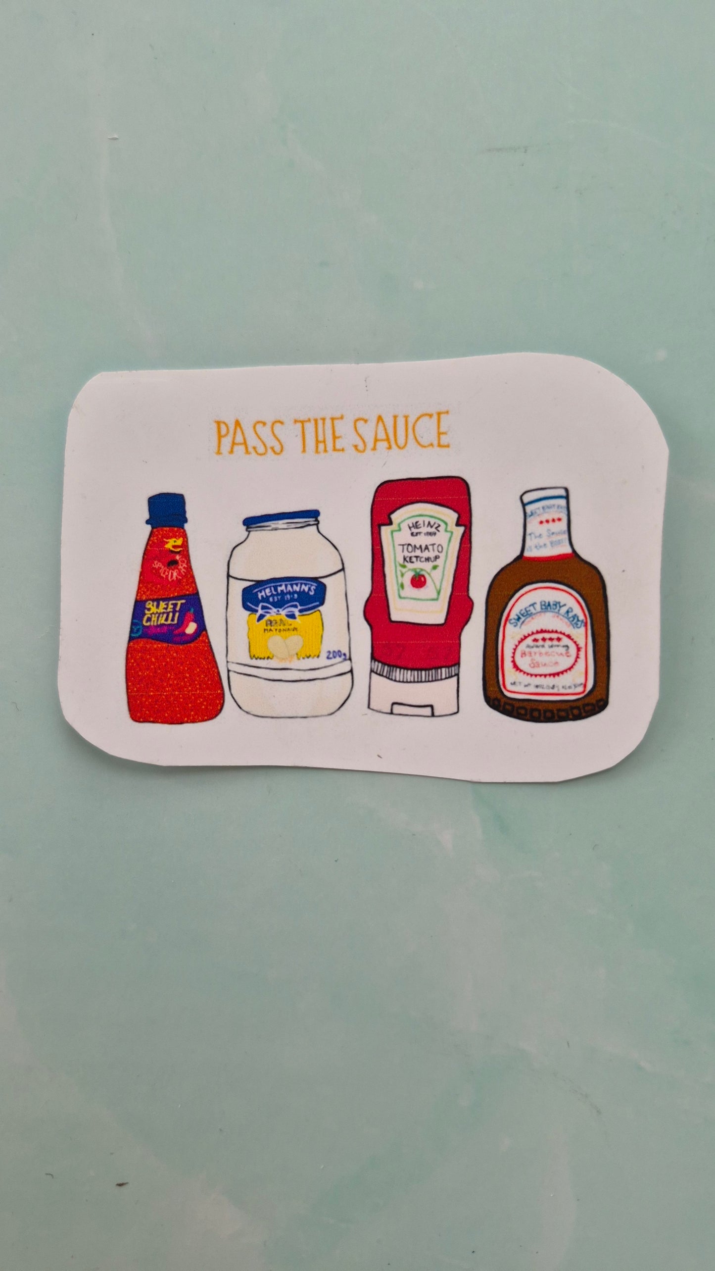 Rectangular vinyl sticker featuring sauce bottle illustrations on a white background and the words ‘PASS THE SAUCE’ above. 