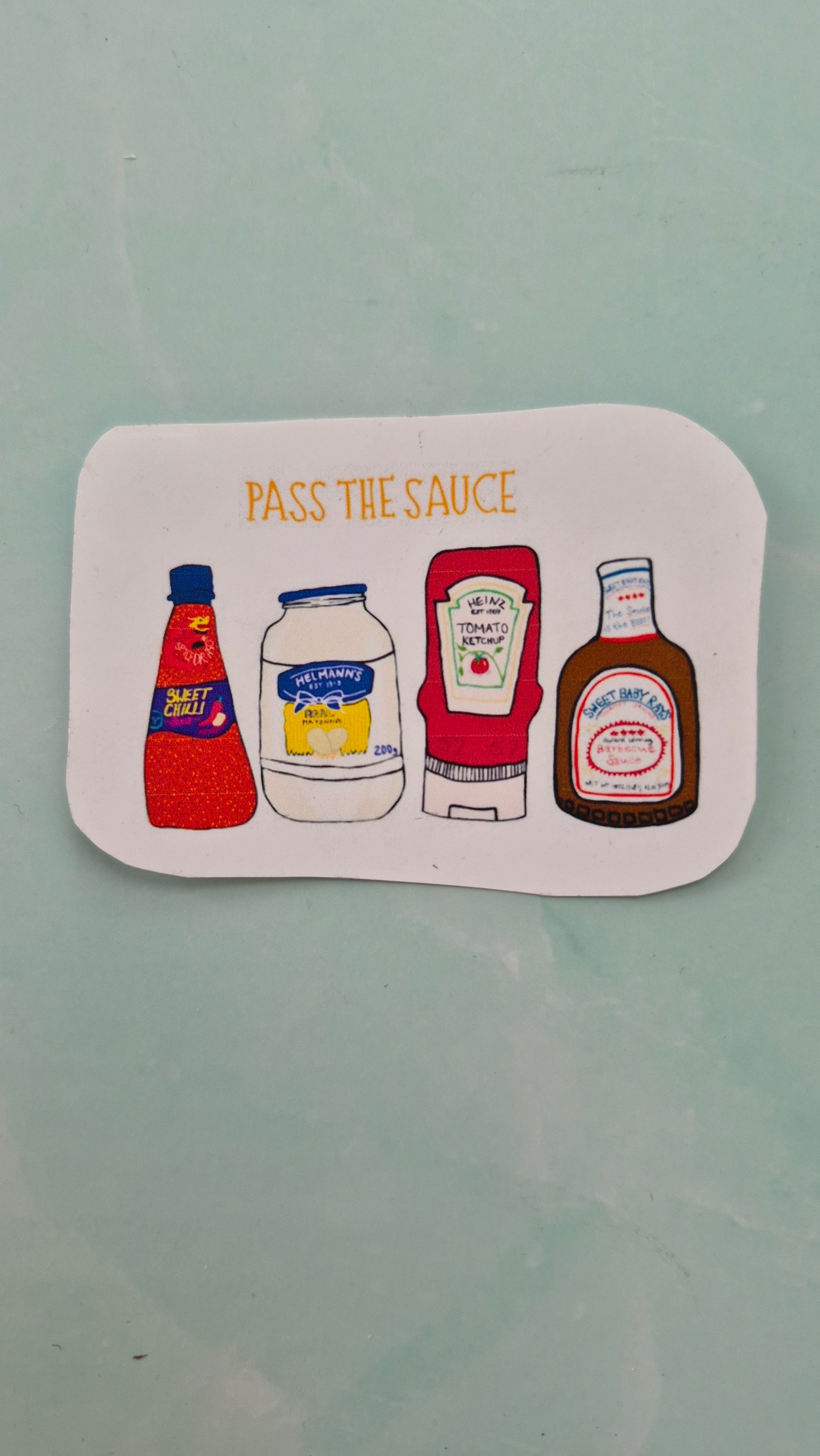 Rectangular vinyl sticker featuring sauce bottle illustrations on a white background and the words ‘PASS THE SAUCE’ above. 