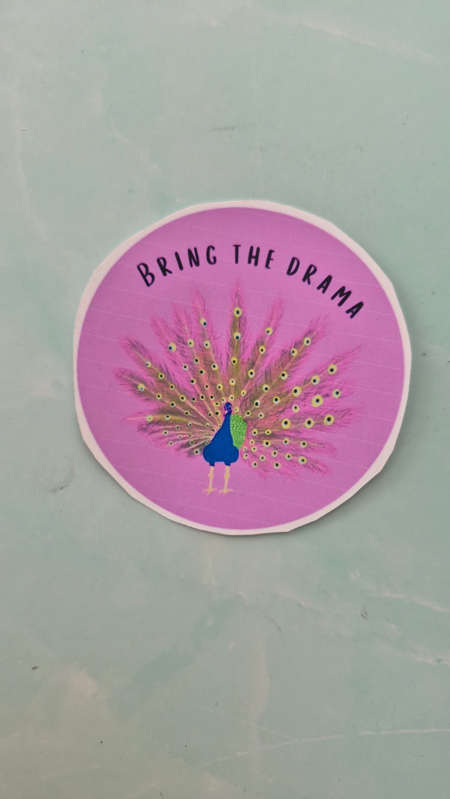 Purple vinyl sticker with. A peacock design and the word ‘bring the drama’ 