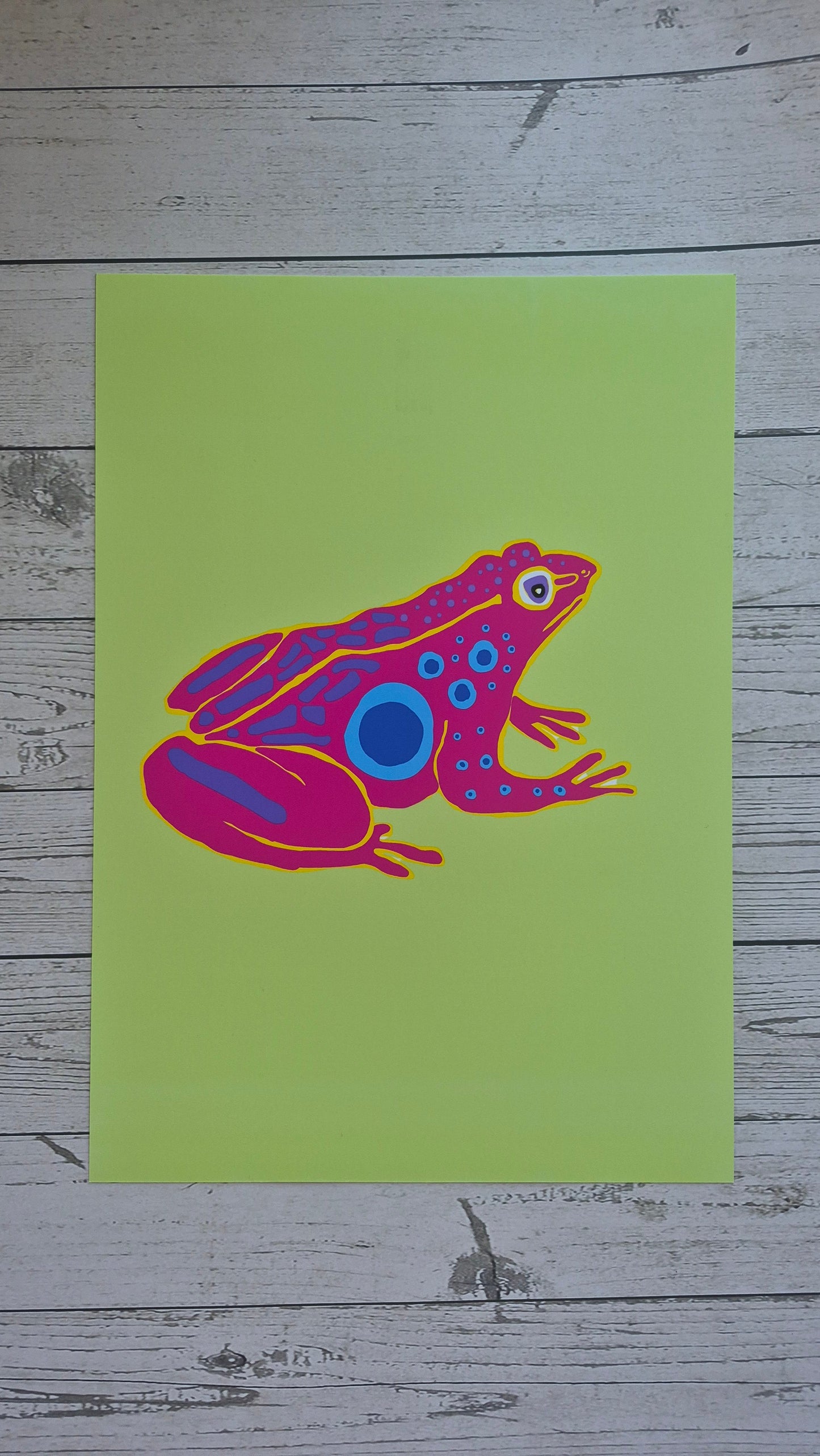 Colourful pink and purple frog illustration on a green card with a wooden background