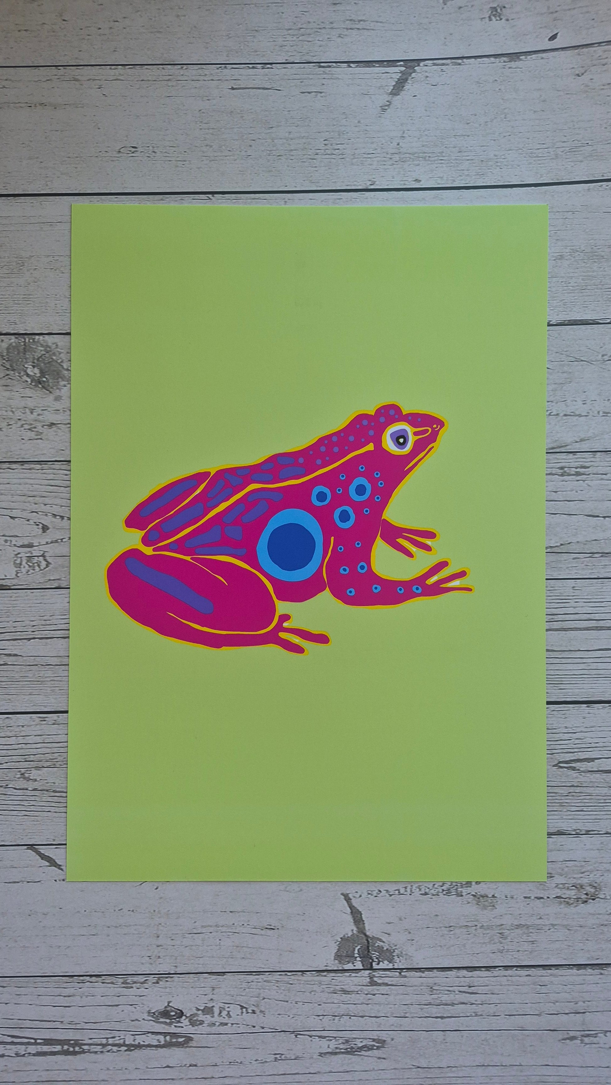 Colourful pink and purple frog illustration on a green card with a wooden background