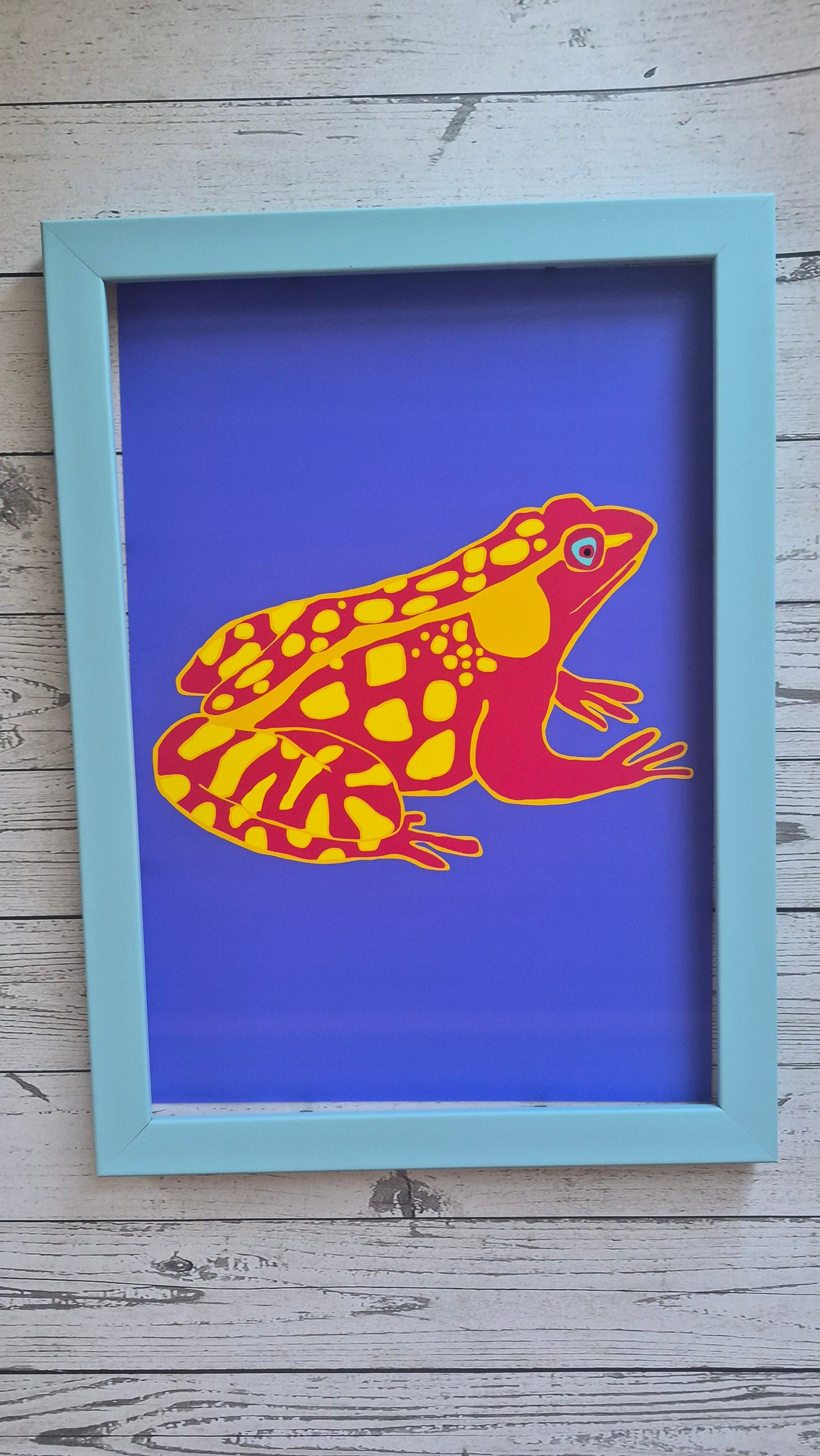 Framed artwork of a colourful abstract red frog design on a wooden surface