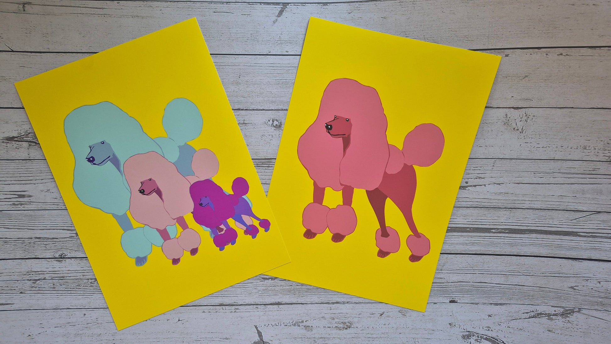 Two colourful poodle illustrations printed on yellow fine art paper against a wooden surface.