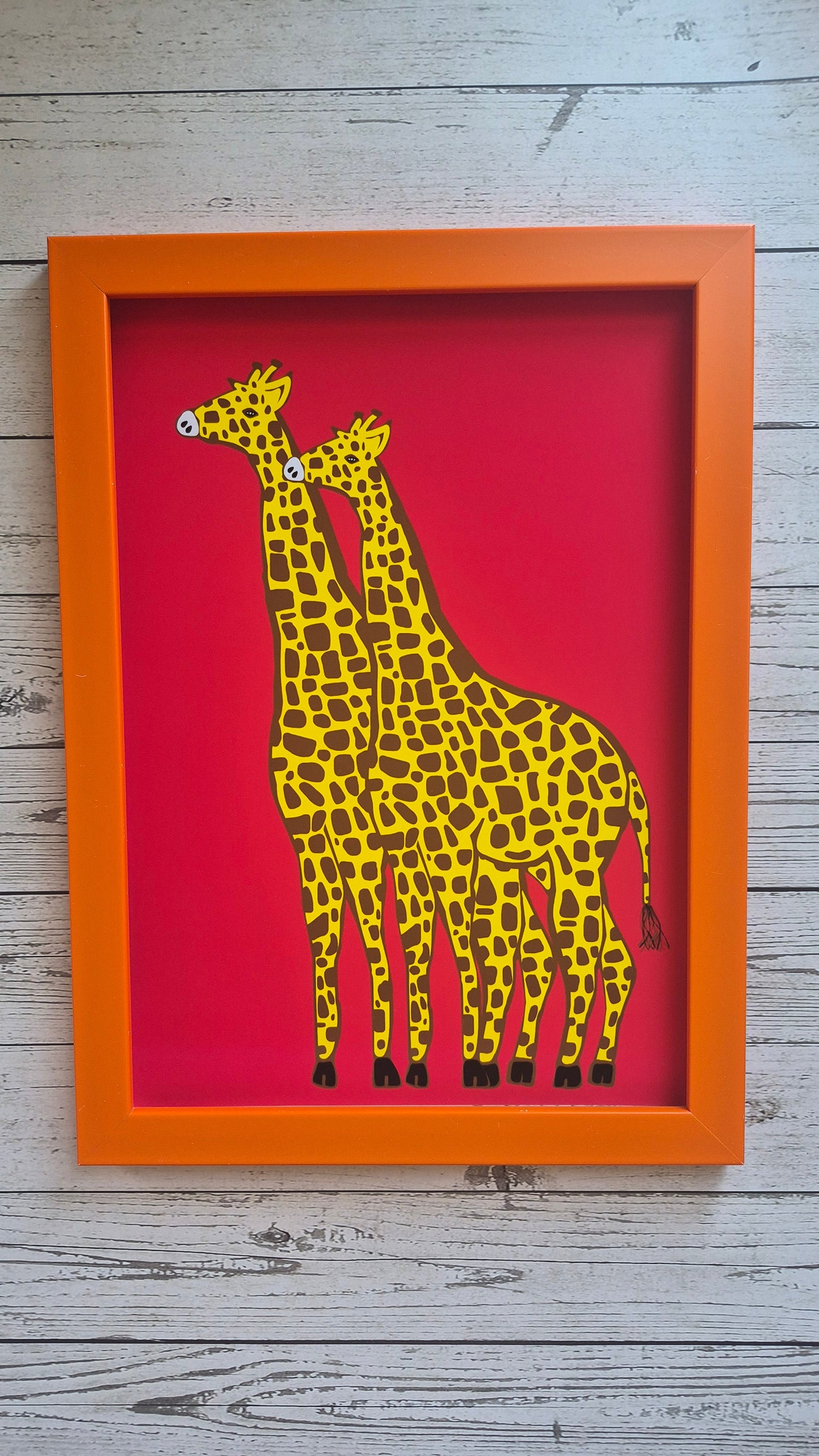 Framed art print featuring two giraffes walking side by side. Solid red background. Orange frame. Placed on an ash wood surface. 
