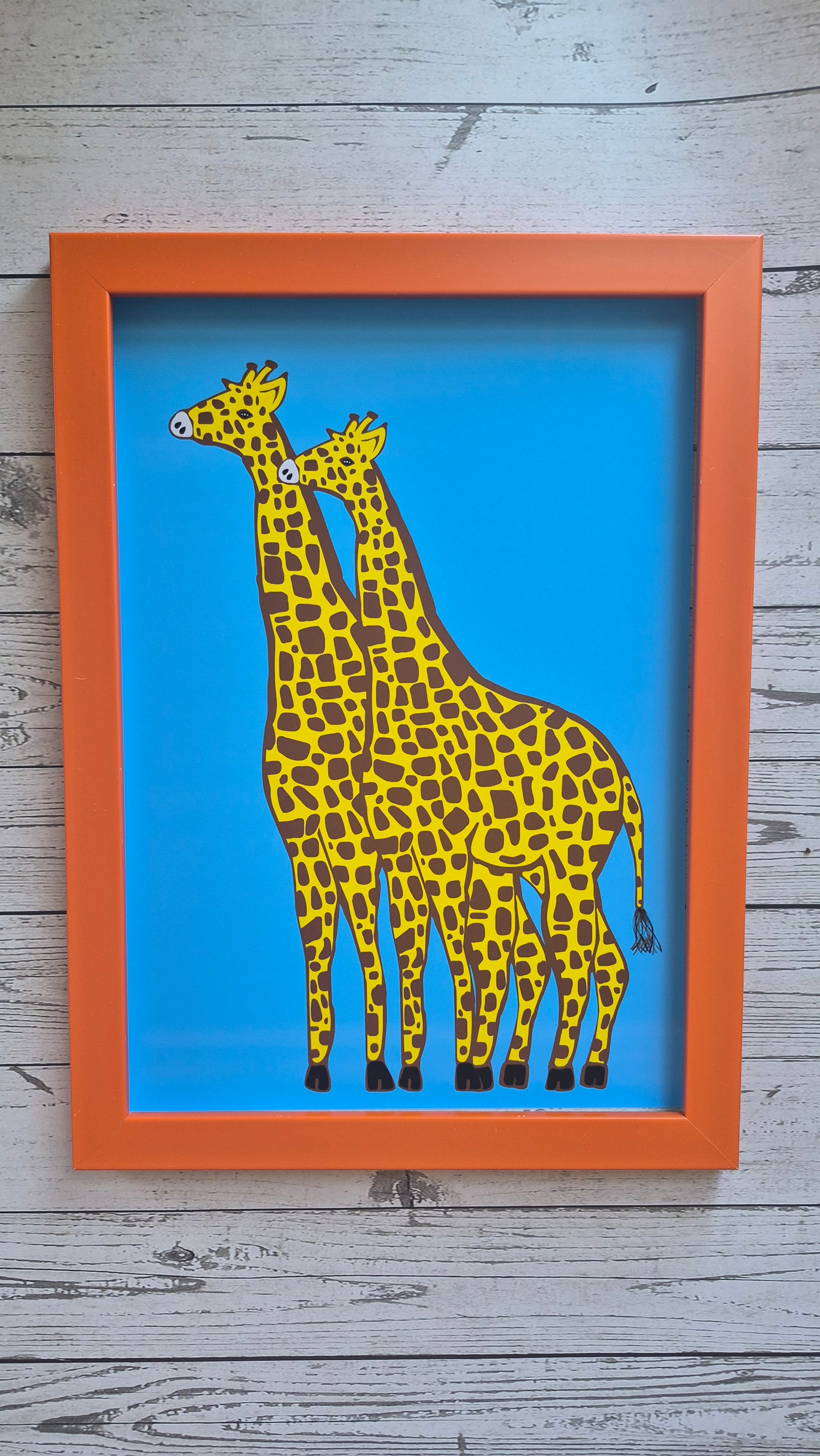 Framed art print of two giraffes on a solid blue background. Framed in an orange frame and resting on a light wooden surface.