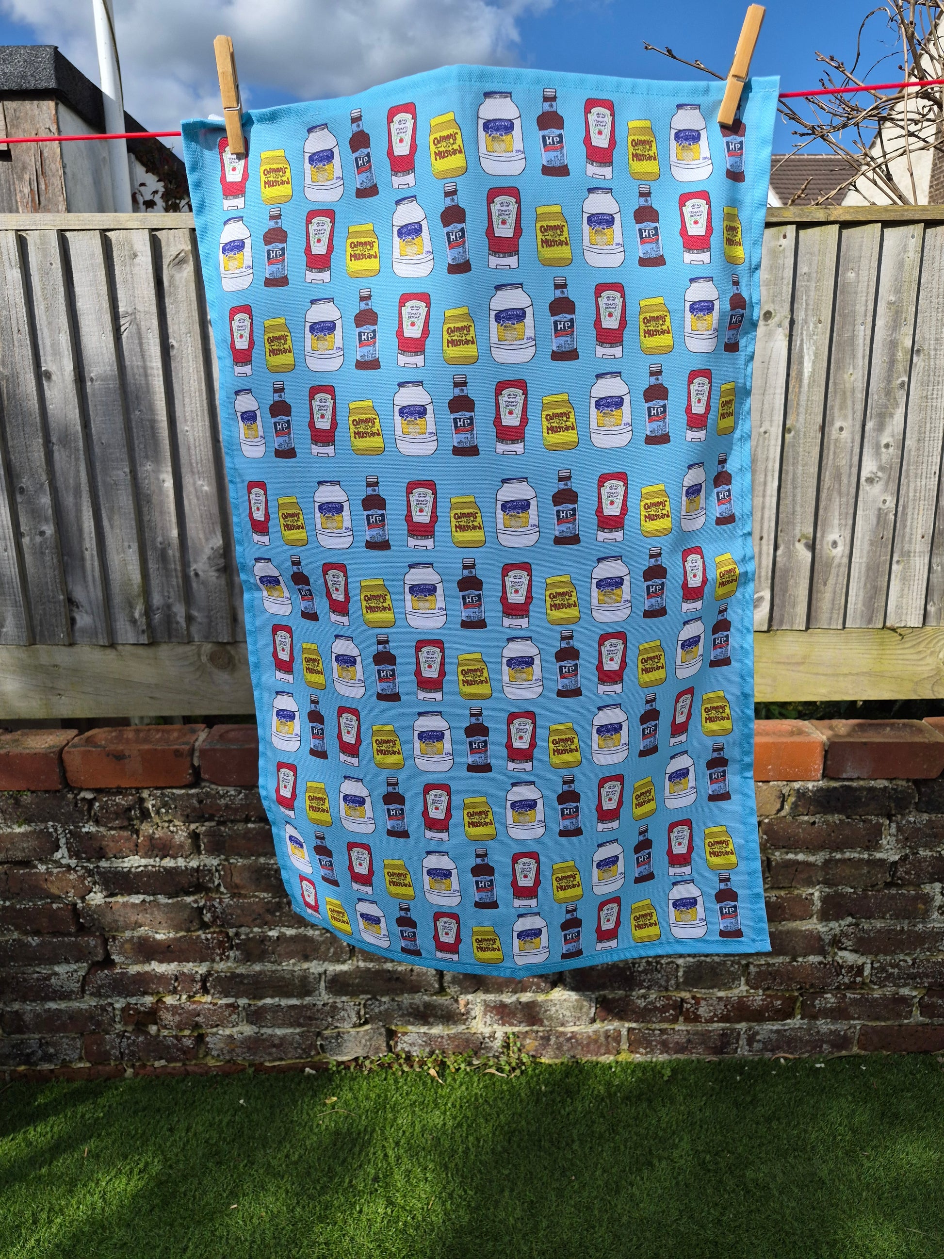 Unique blue tea towel with hand drawn condiments print hanging on a washing line. 