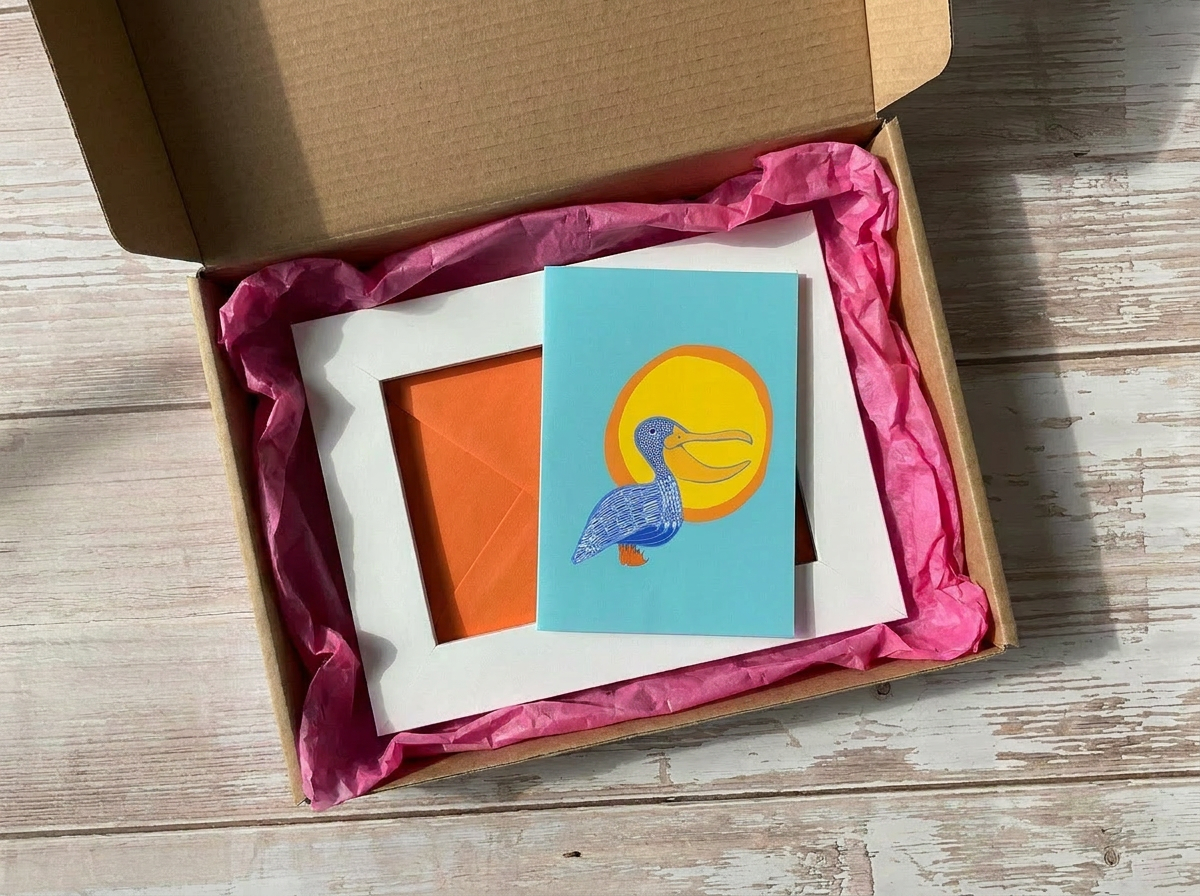 Open box with pink tissue paper containing a card with a pelican design on a wooden surface