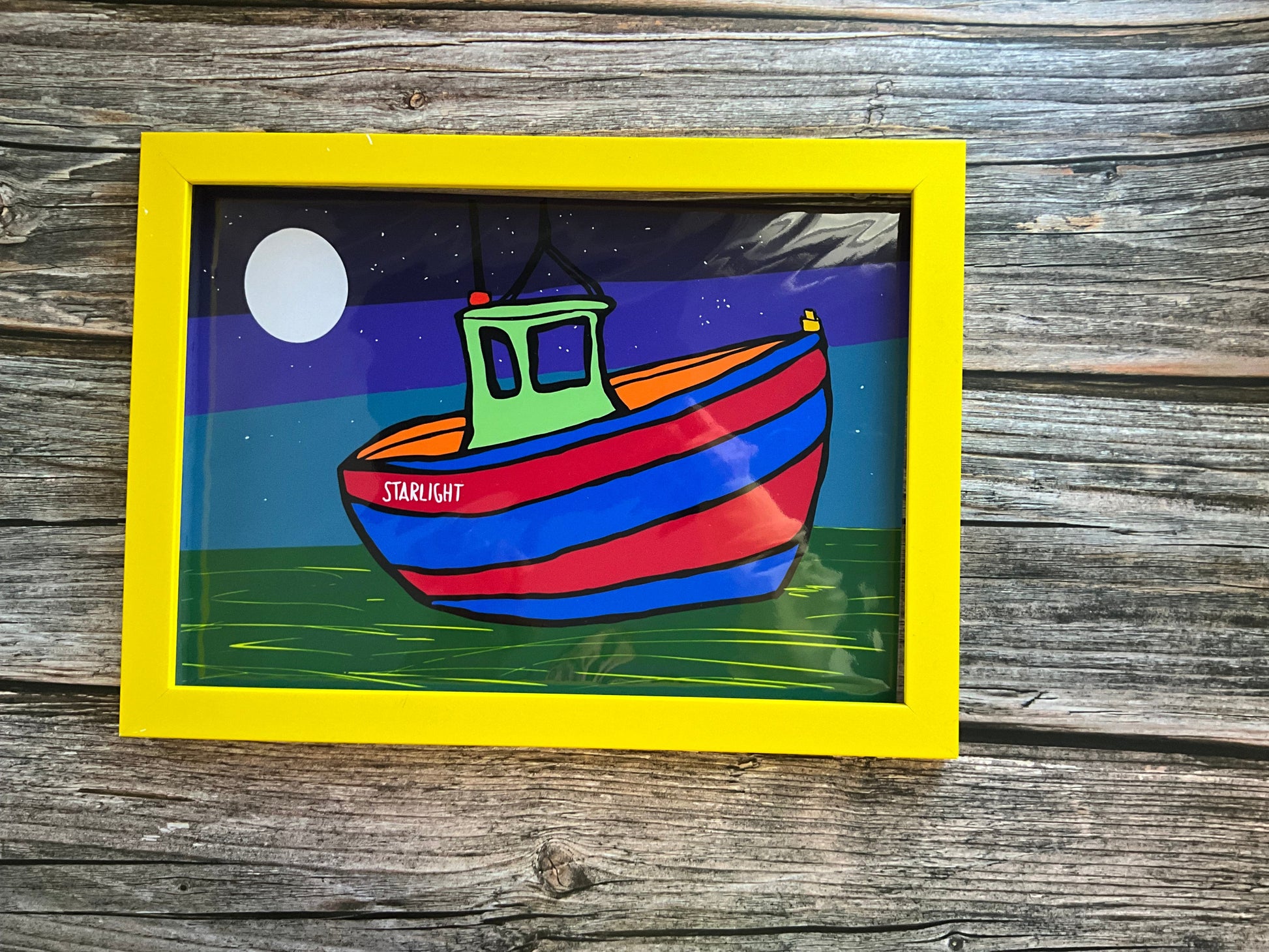 Colourful sail boat art print with a moon and starry sky. Yellow frame on a wooden surface. 