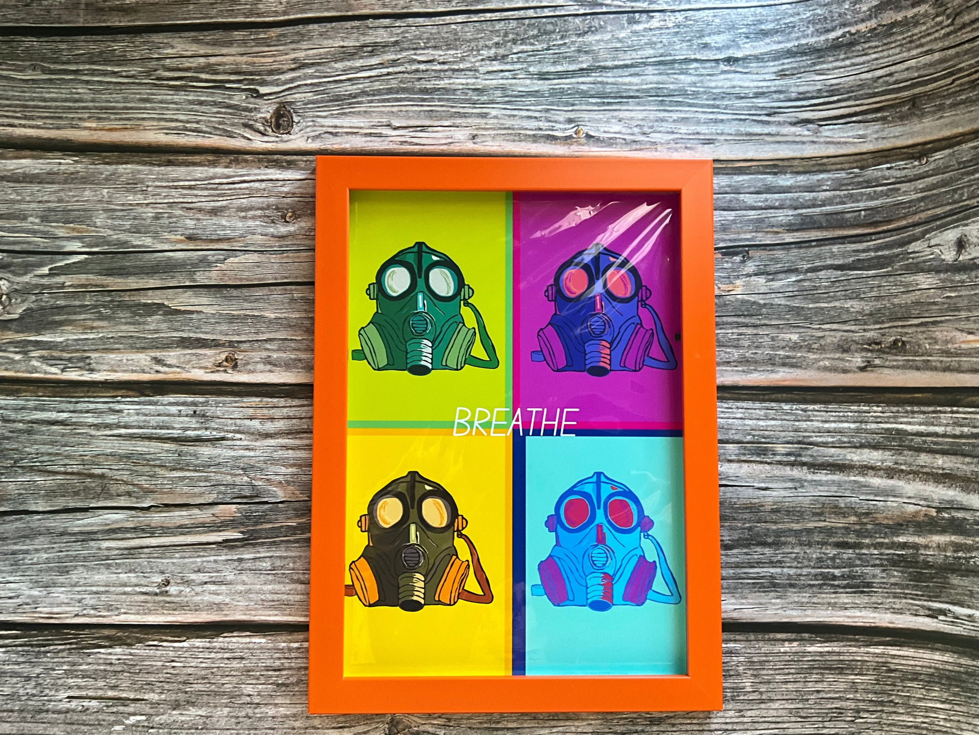Framed artwork print with colourful gas mask designs and the word ‘BREATHE’ in white letters. The print is placed  on a wooden background