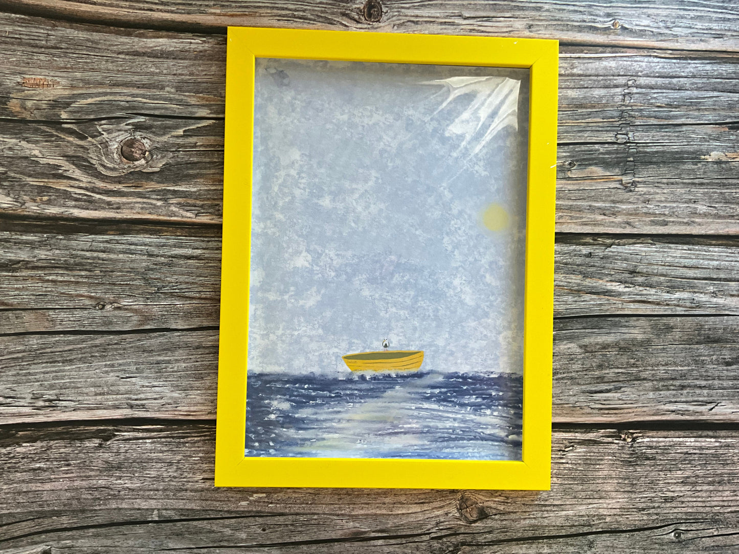 Framed artwork of a boat on water with a seagull perched on it, framed in a yellow frame border on a wooden surface.