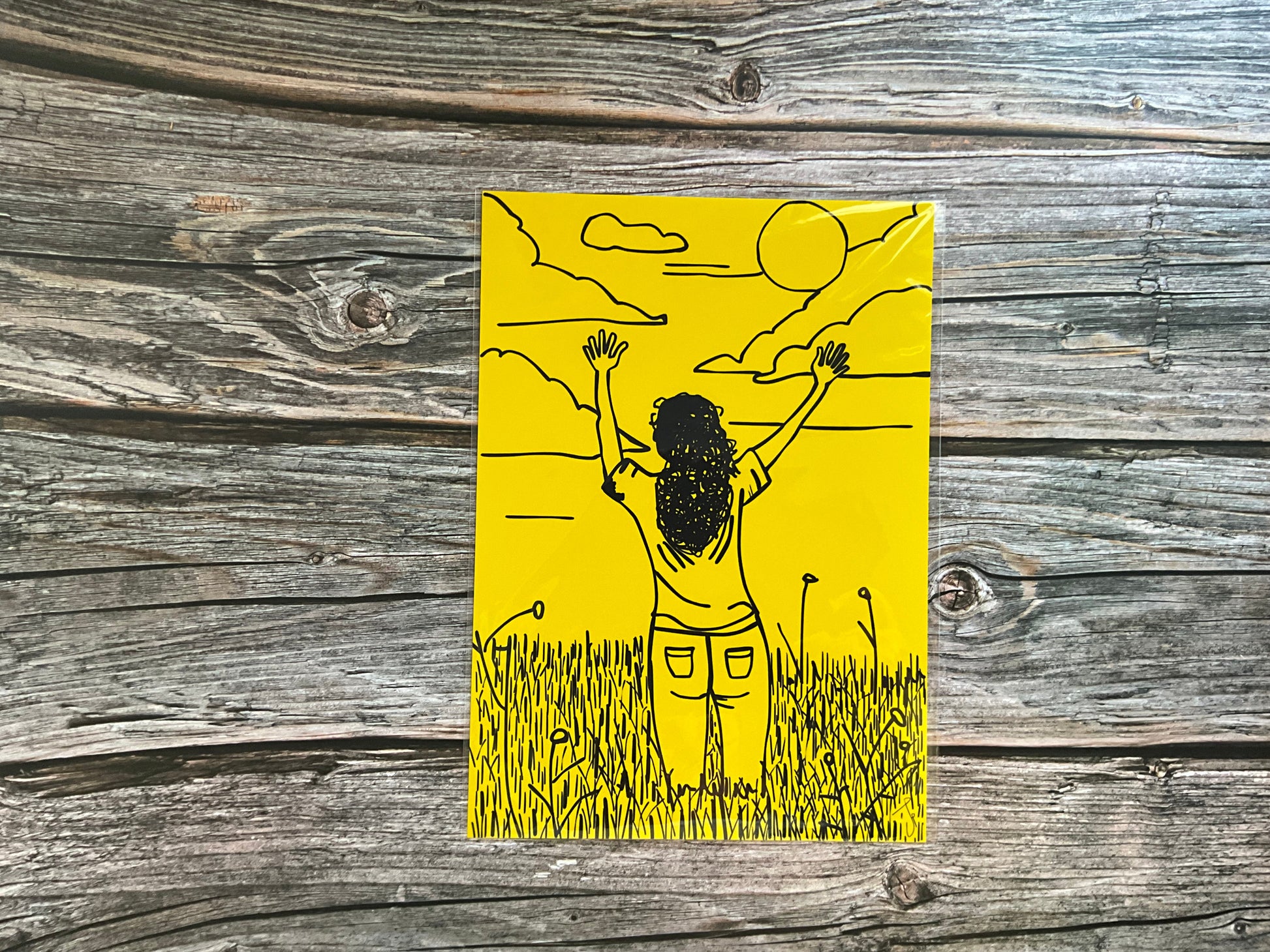 Yellow outline illustration of a girl with arms outstretched  to the sky. The print has a bright yellow background and is placed ion a wooden surface.