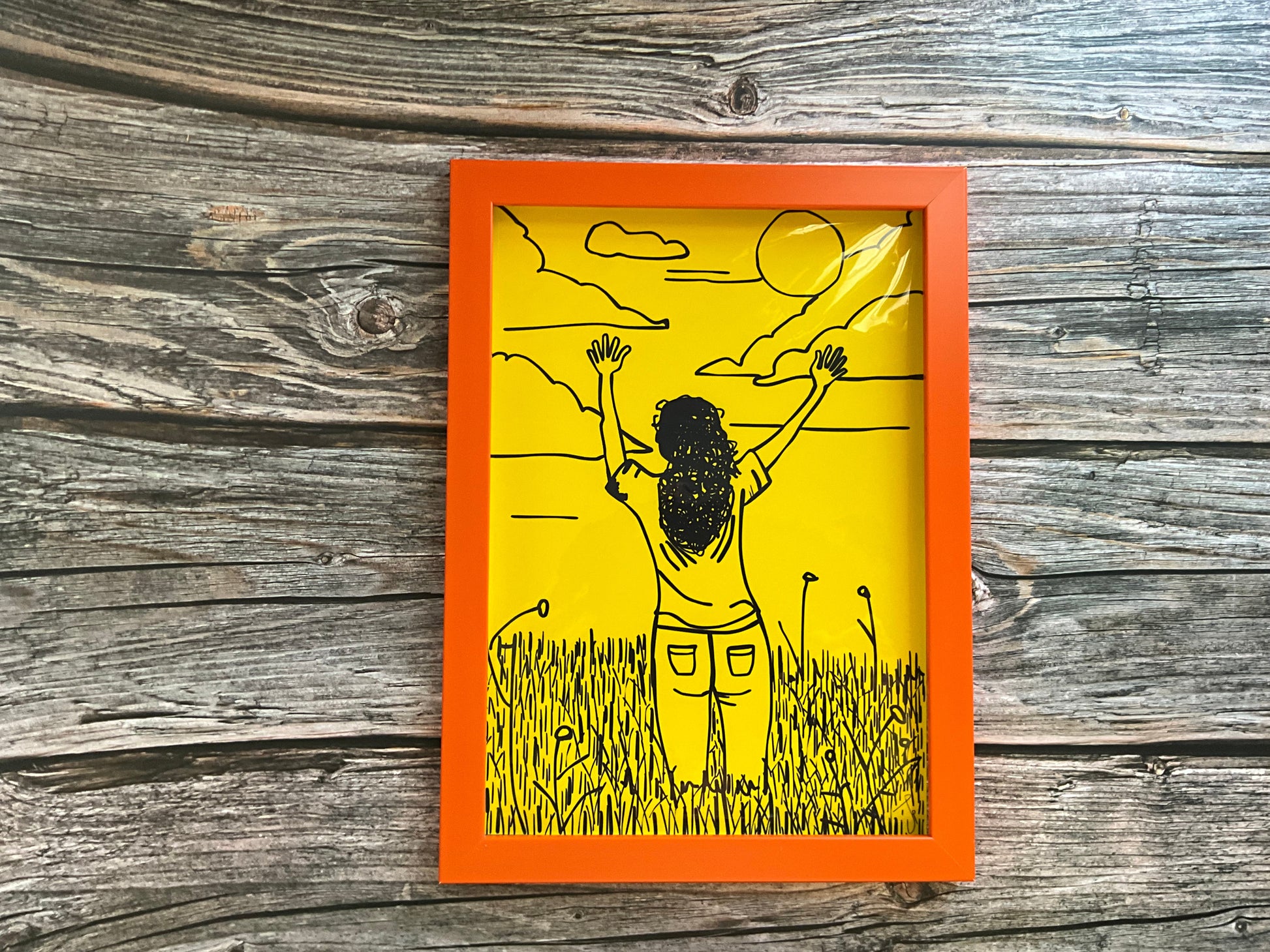 Art print of a girl in a meadow field raising her hands to the sky. The print is framed in an orange frame and has a bright yellow background. 