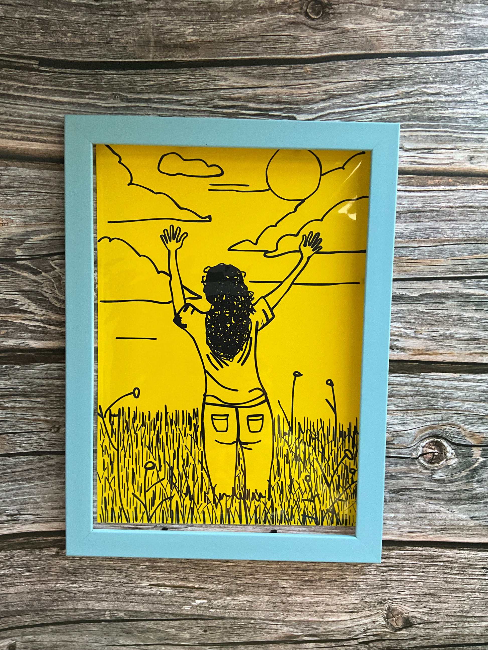 Art print of a girl in a meadow field raising her hands to the sky. The print is framed in an blue frame and has a bright yellow background. 
