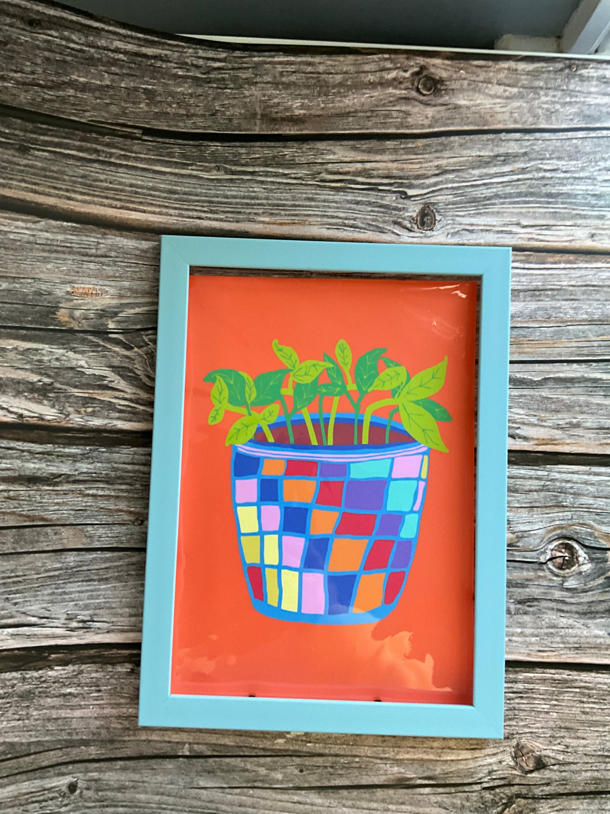Framed artwork with a colourful pot plant with green leafy plant on an orange background, mounted on a wooden wall.