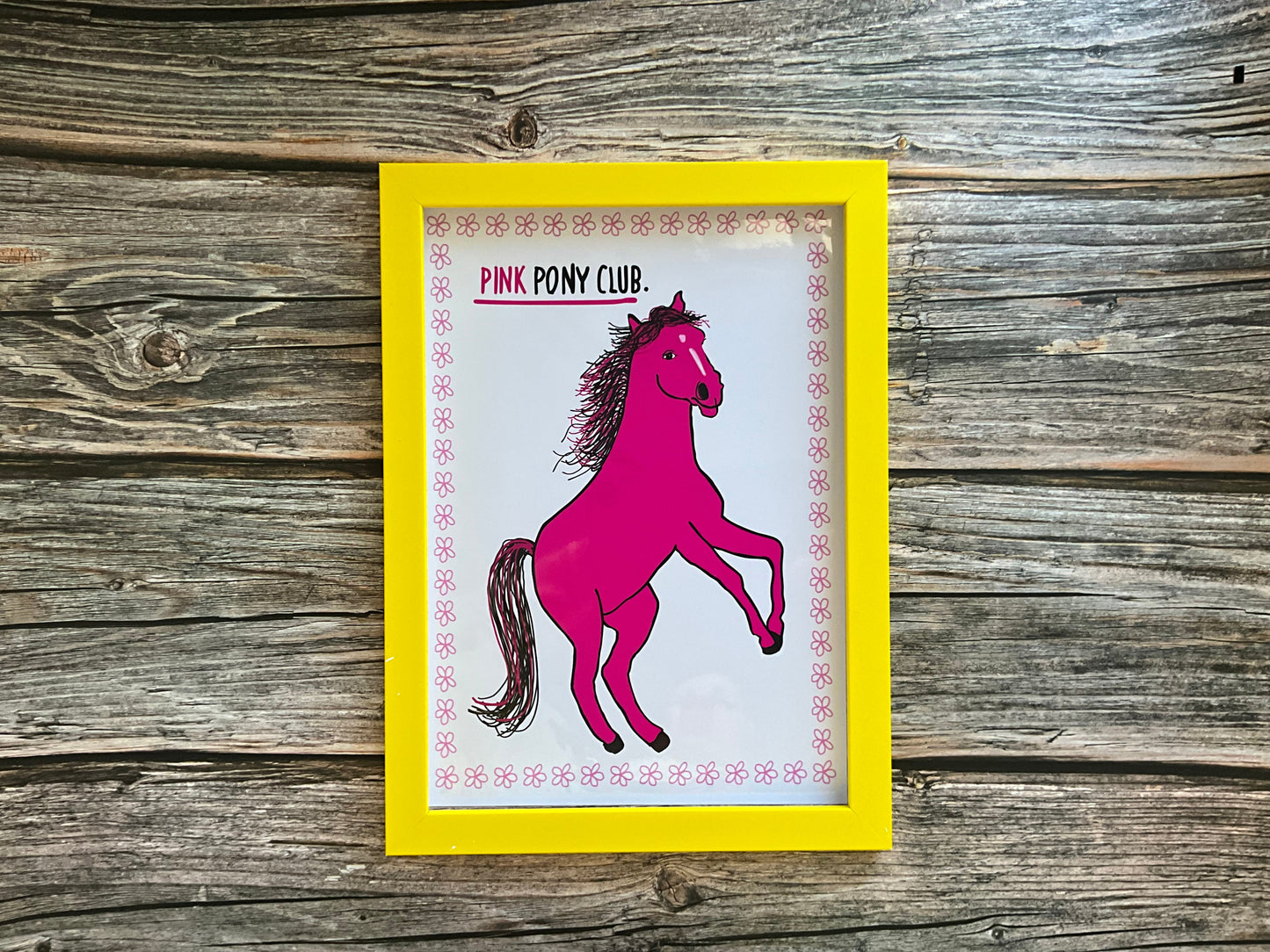 Pink horse print with the text ‘pink pony club’ White background with a pink flower outline. The print is in a yellow frame resting on a wooden surface. 