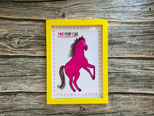 Pink horse print with the text ‘pink pony club’ White background with a pink flower outline. The print is in a yellow frame resting on a wooden surface. 