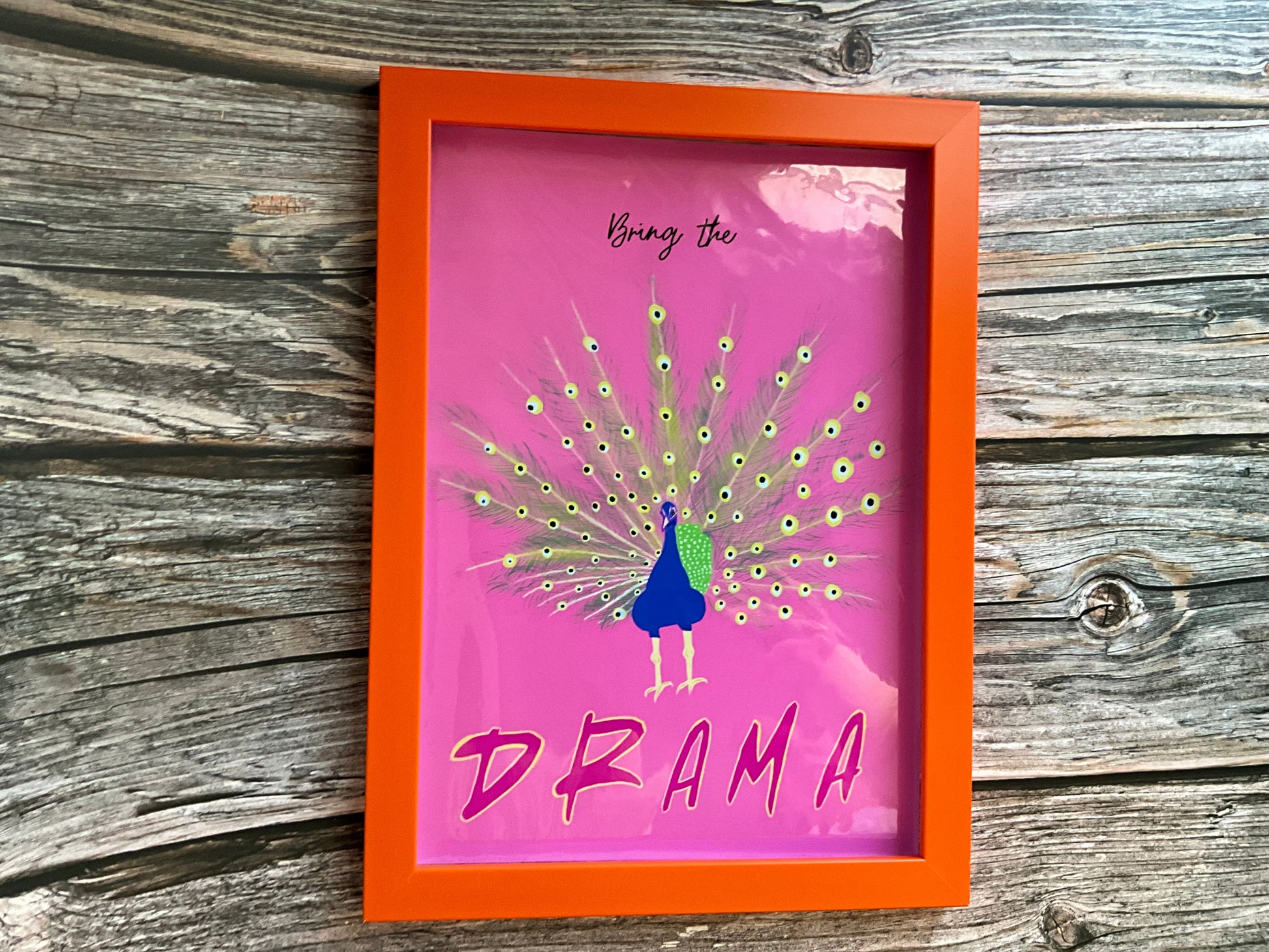 Framed artwork with a peacock illustration and text 'Bring the Drama' on a pink background, placed on a wooden surface.