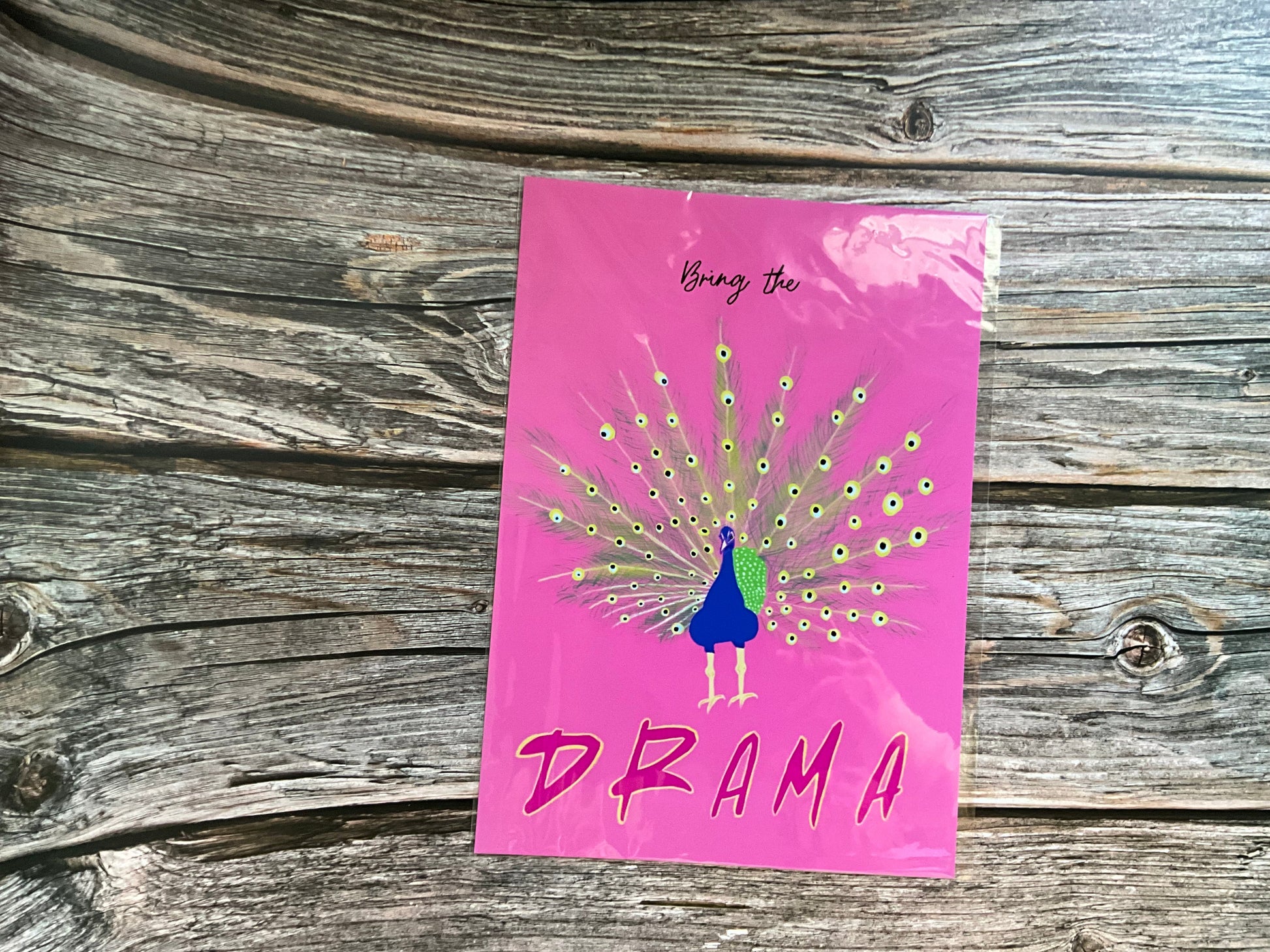 Pink art print with a peacock illustration and 'Bring the Drama' text on a wooden surface