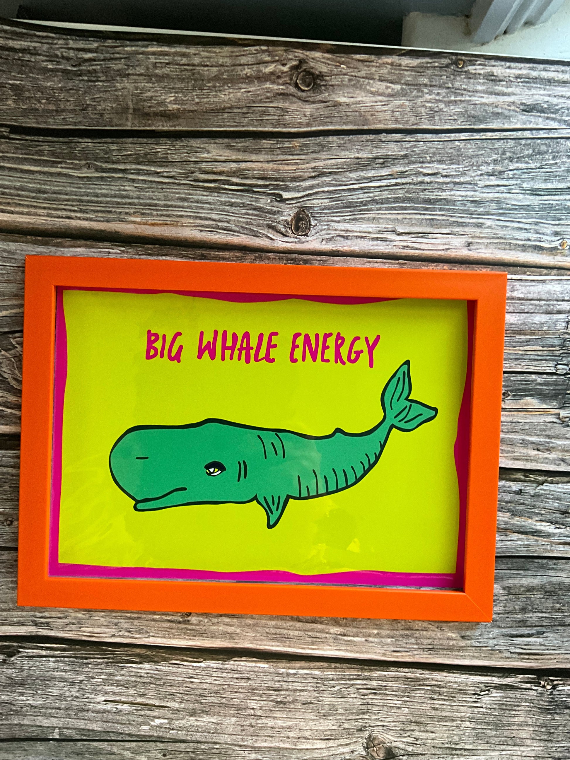 Framed artwork with a green whale illustration and 'Big Whale Energy' text on a wooden surface.