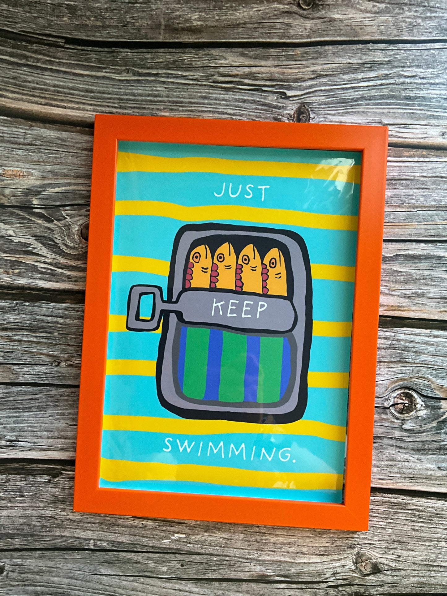 Framed in an orange frame. Mackerel wall art print featuring colourful mackerel fish in a green and blue tstriped it in on a yellow and blue striped background. The words ‘Just keep swimming are on the tin’
