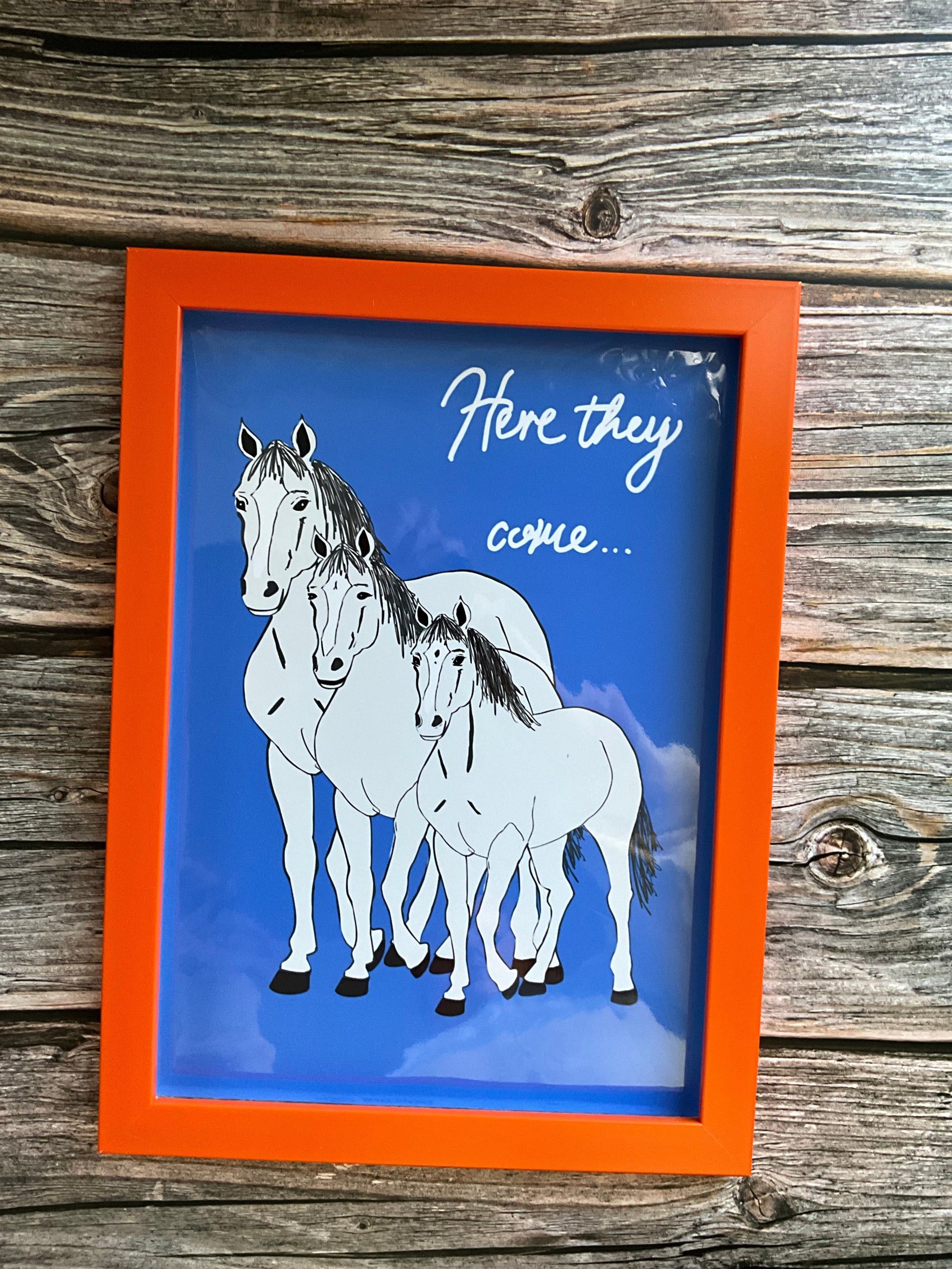 Framed artwork of a horse on a blue background with an orange border, displayed on a wooden surface.