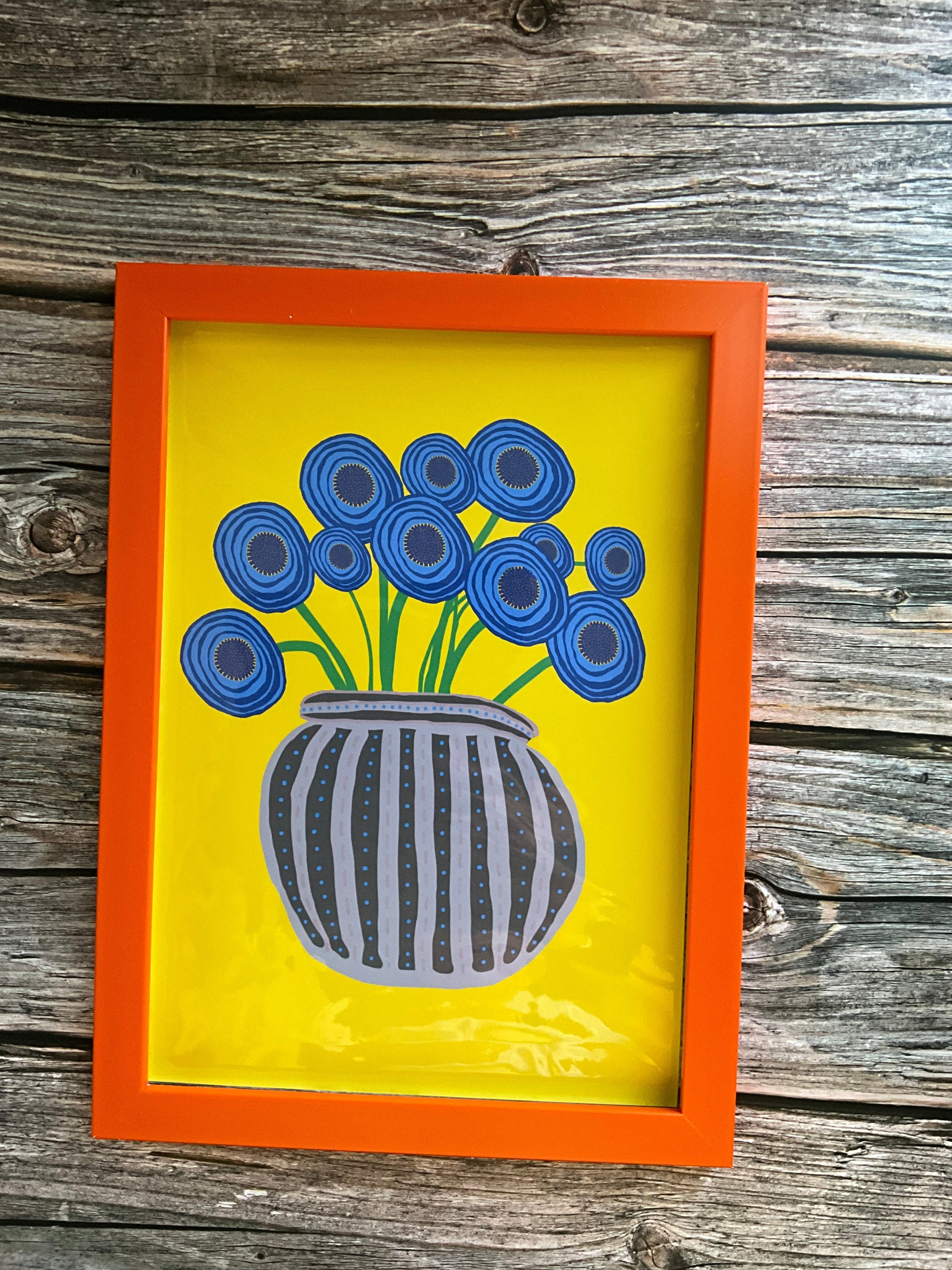 Abstract blue poppy art print with a yellow background and an orange frame, placed on a wooden background