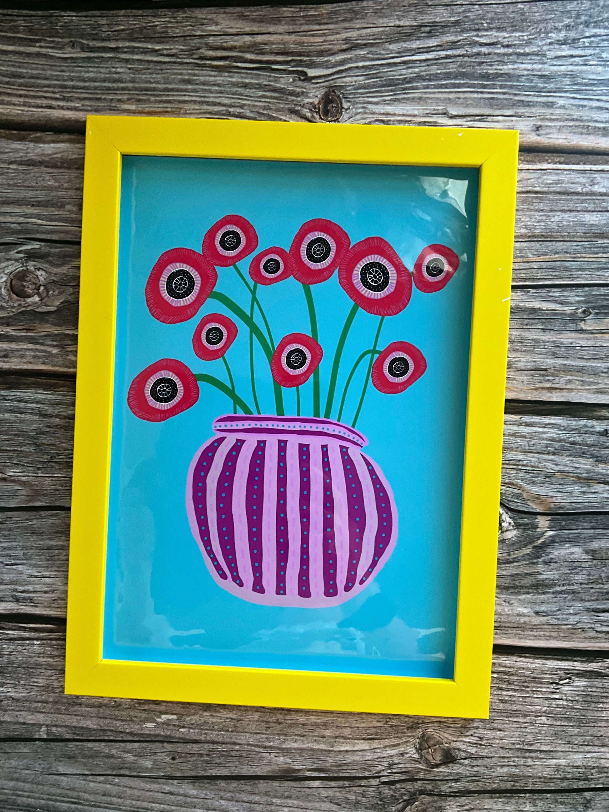 Abstract red poppies in a striped vase. Art print in yellow frame on a wooden background.