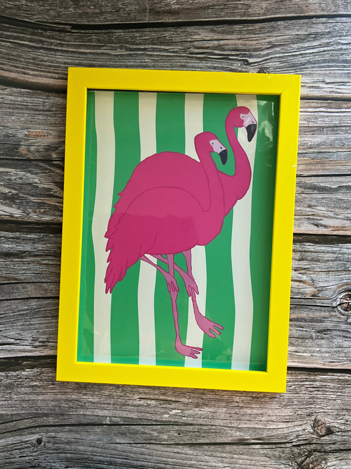 Framed artwork of a pink flamingo on green and white striped background, placed on a wooden surface.