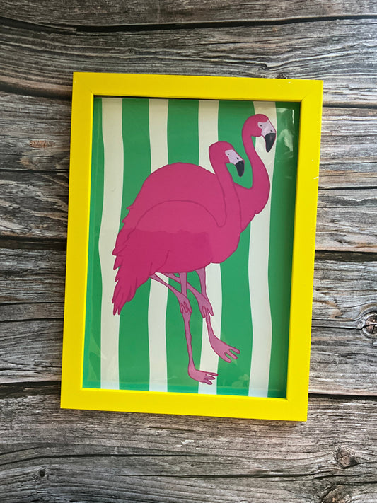 Framed artwork of a pink flamingo on green and white striped background, placed on a wooden surface.