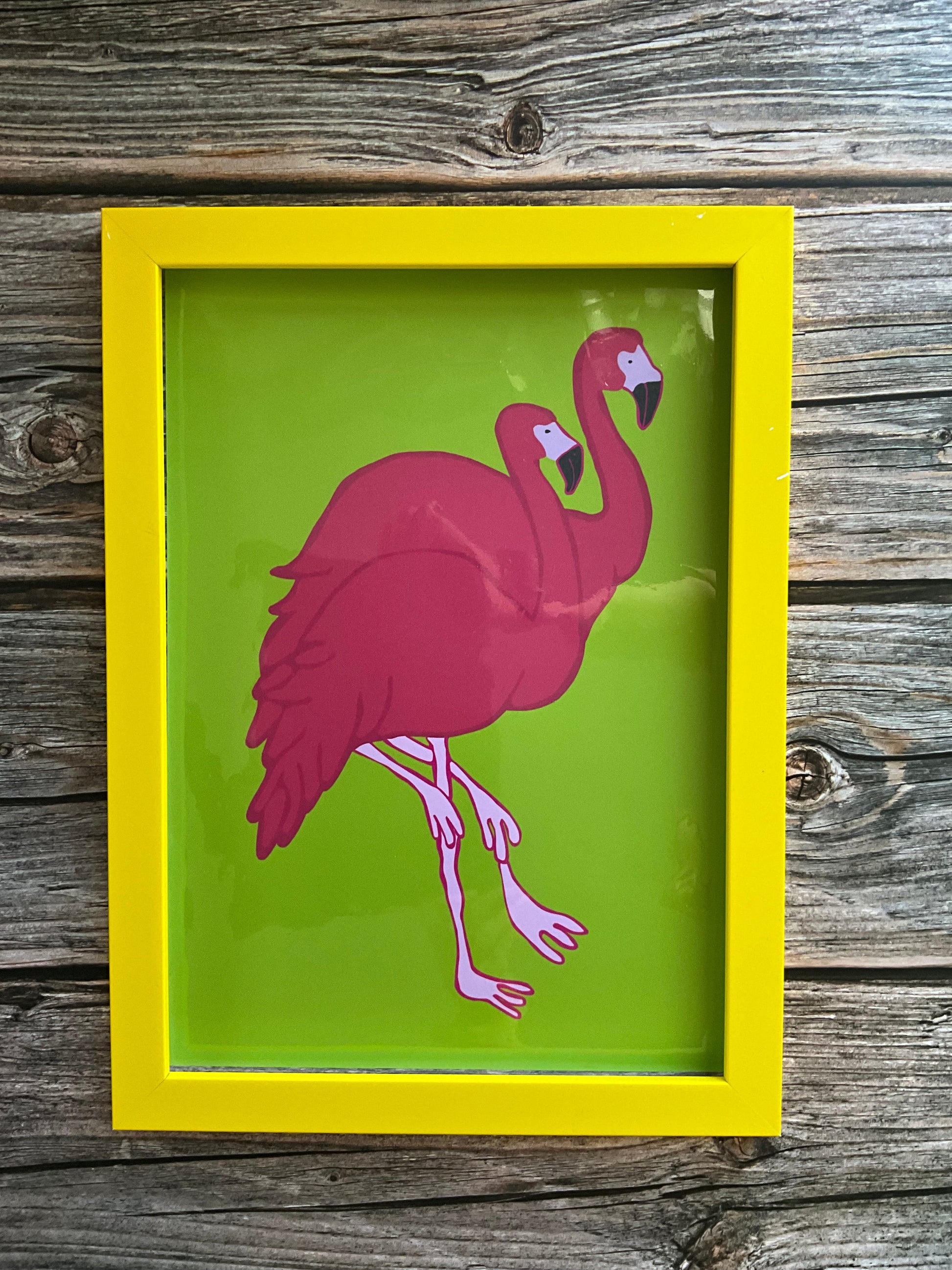 Framed artwork of a pink flamingo on a green background, set against a wooden wall.