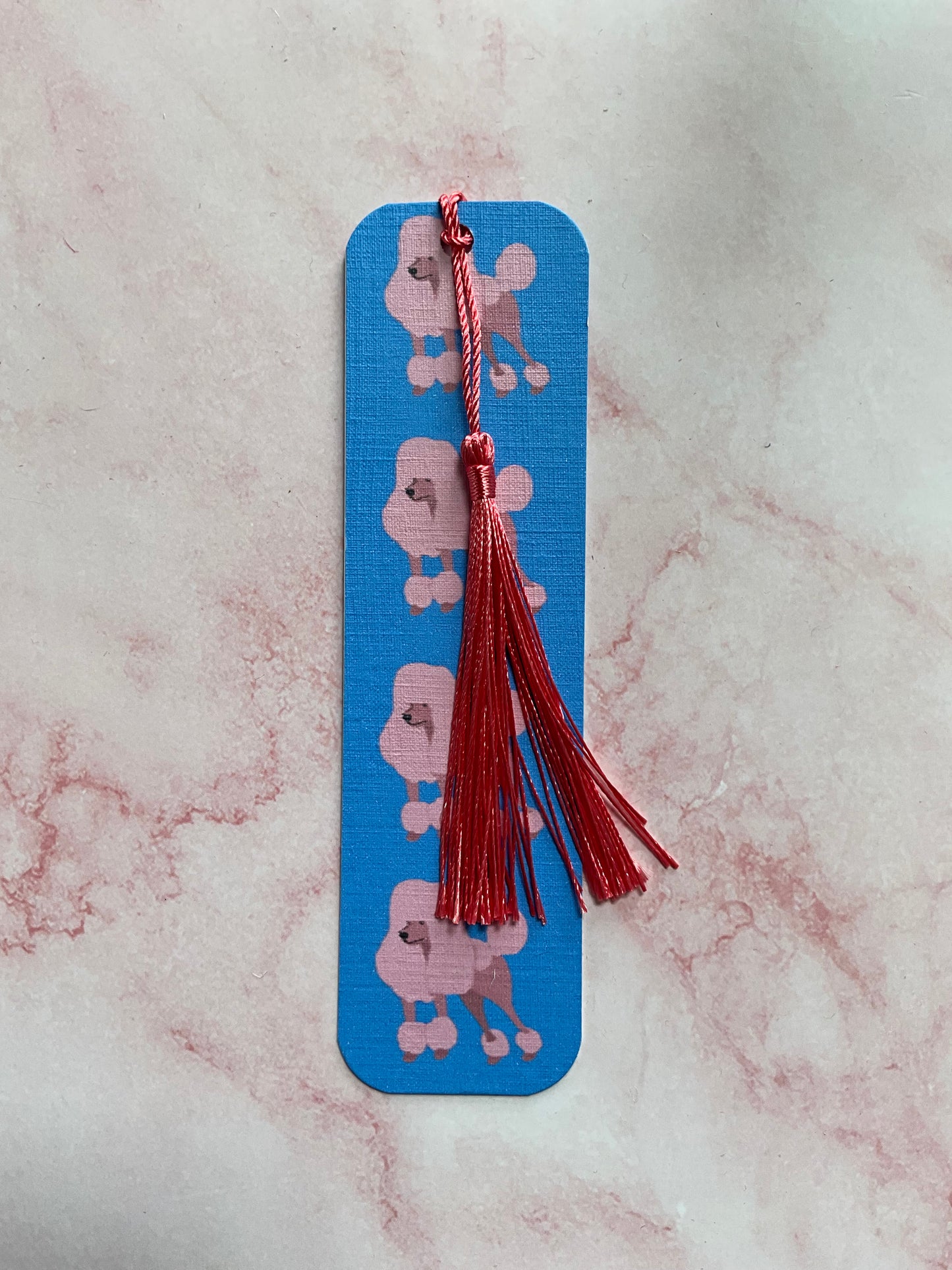 Blue bookmark with pink poodle prints and a pink tassel on a pink marble surfae