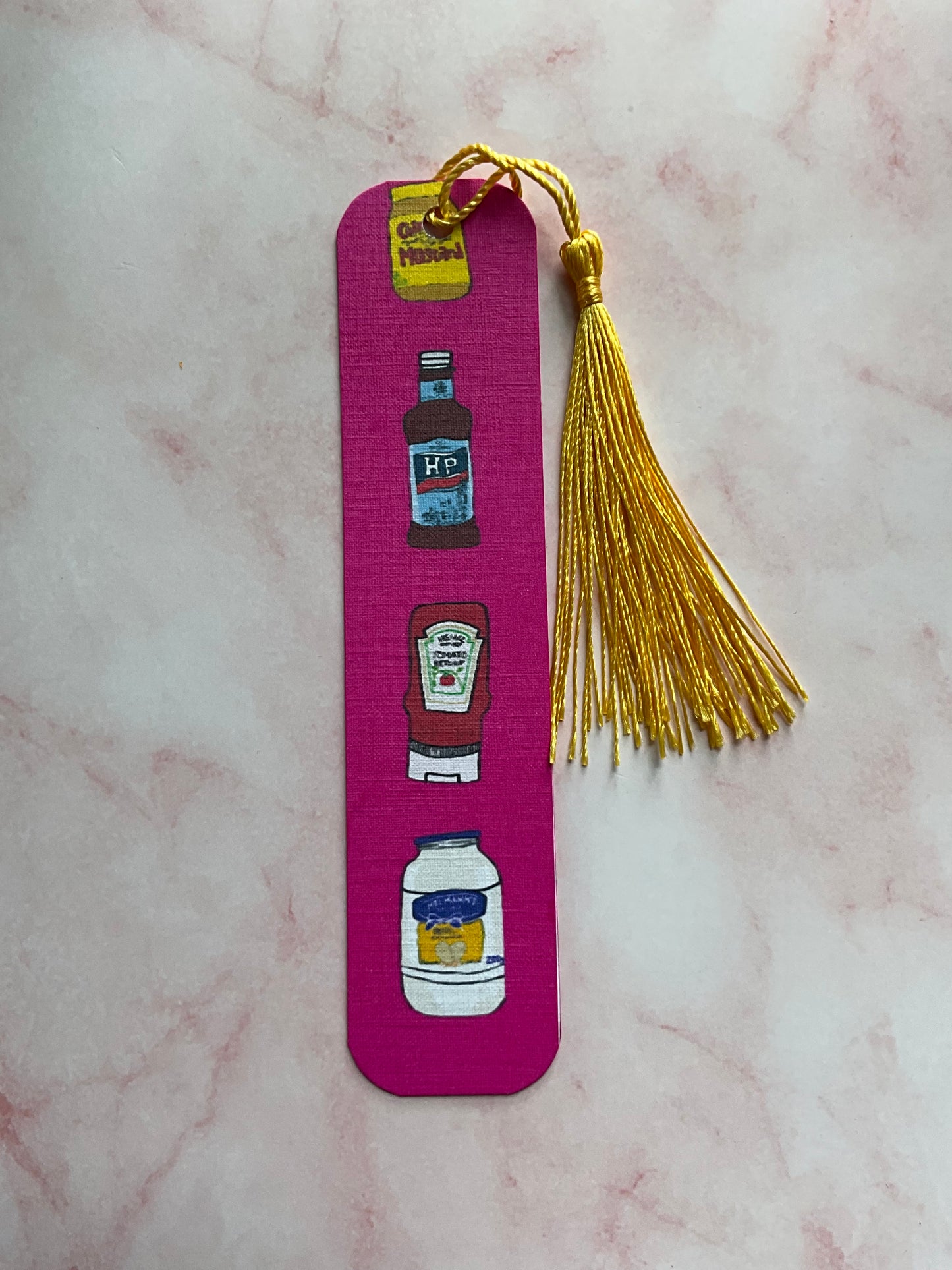 Pink bookmark with condiment illustrations and a gold tassel on a marble surface