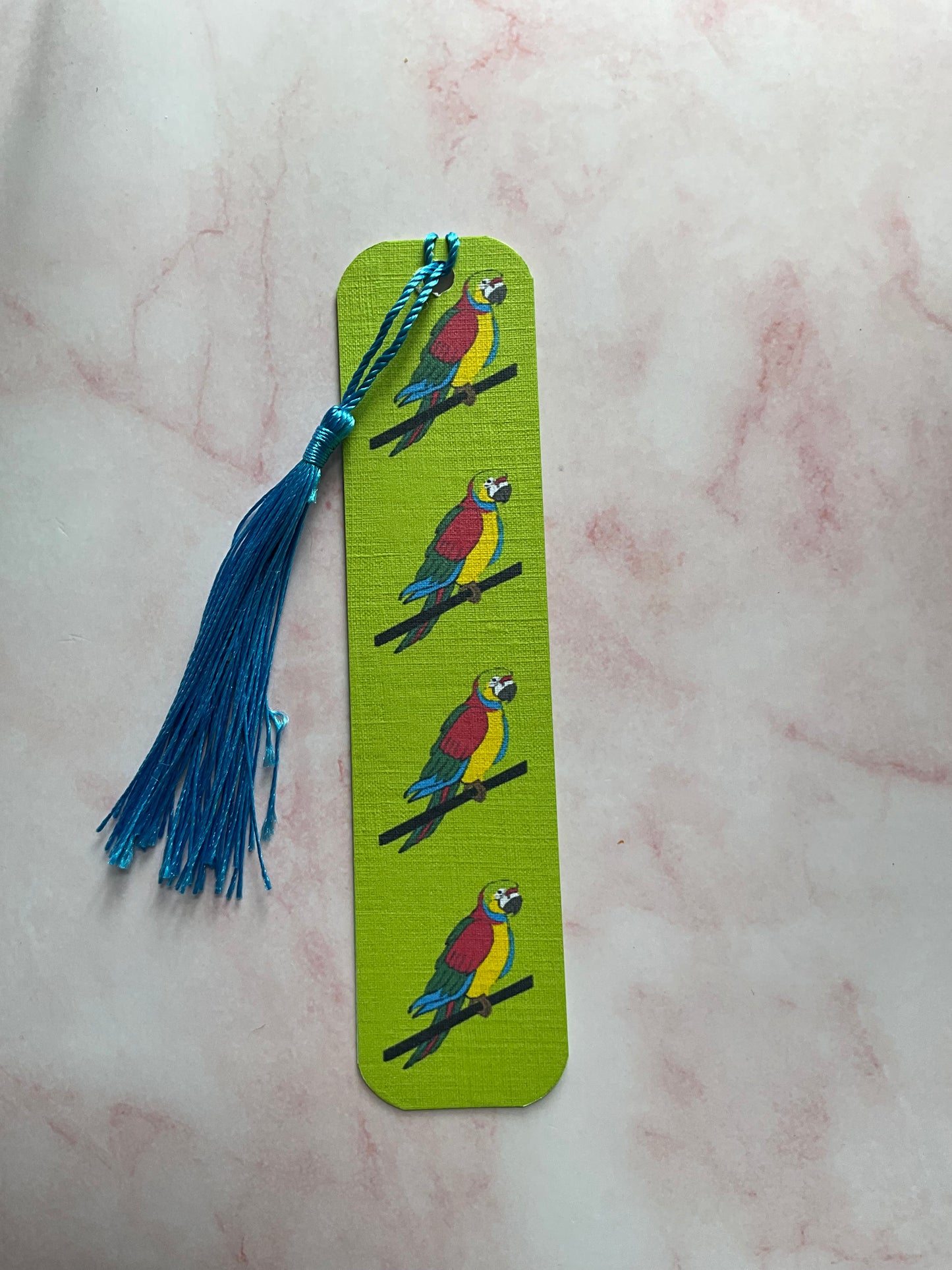 Lime green bookmark with parrot illustrations and a blue tassel on a light pink marble surface
