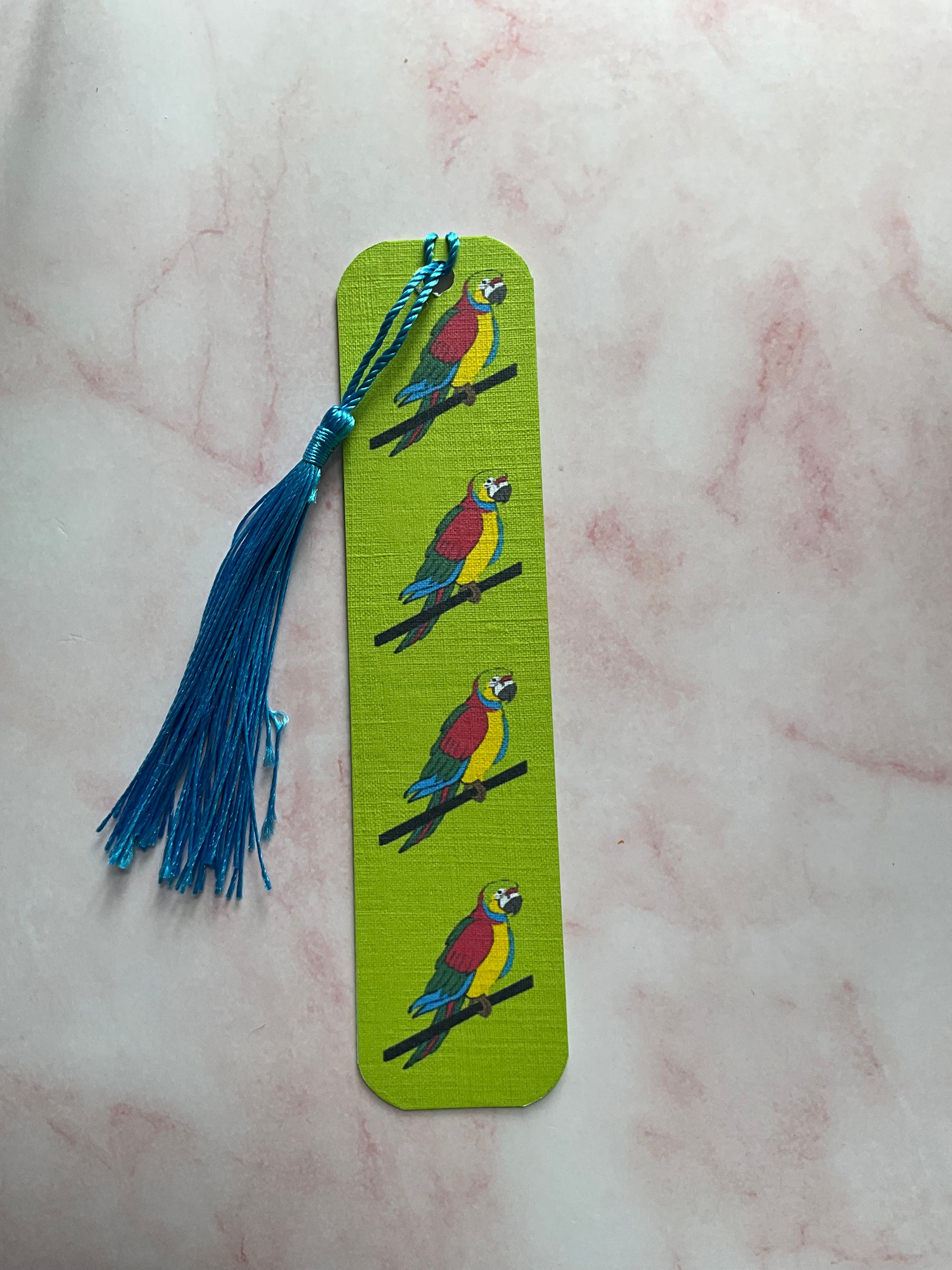 Lime green bookmark with parrot illustrations and a blue tassel on a light pink marble surface