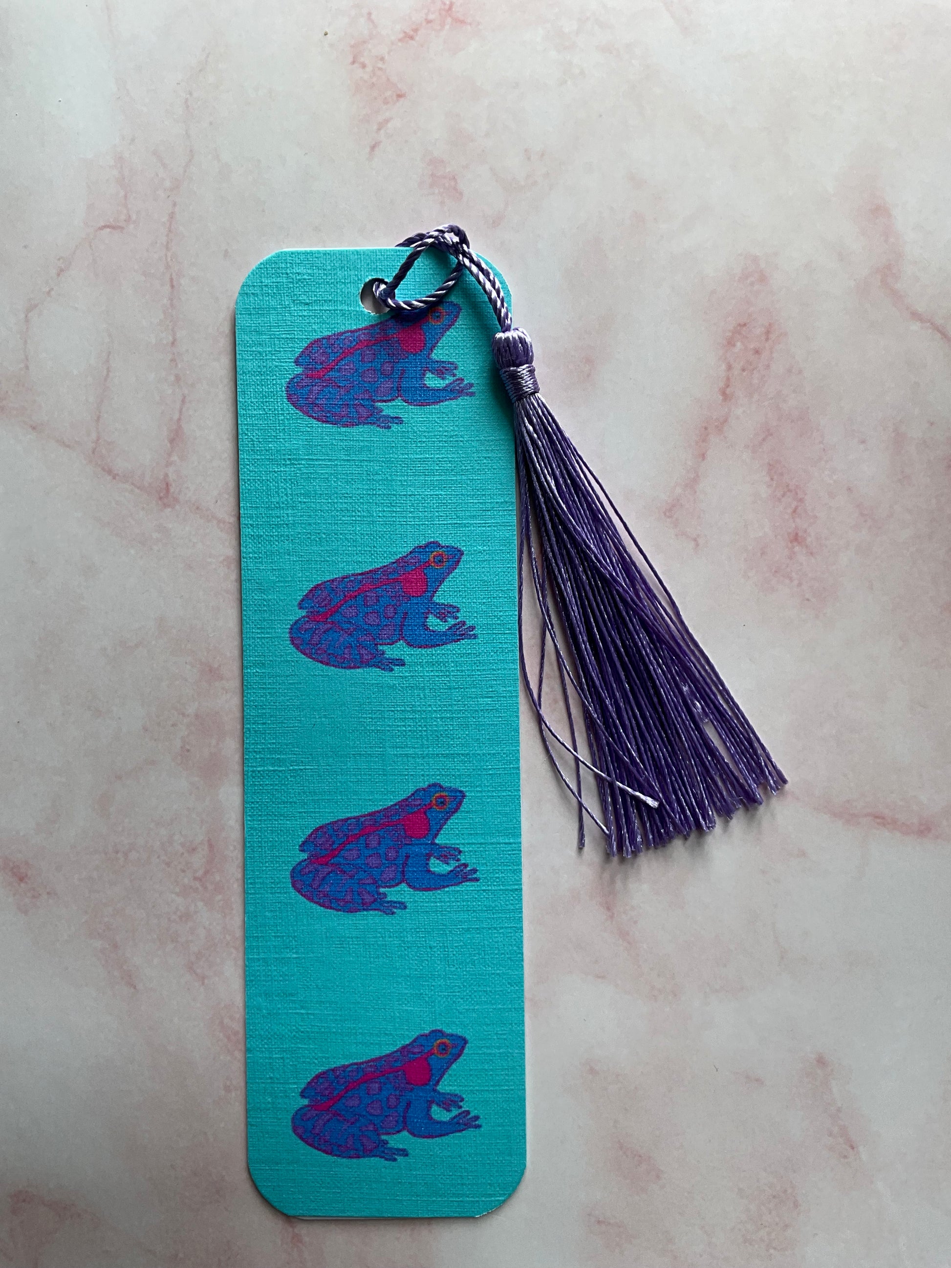 Blue bookmark with purple frog design and purple tassel on a pink marble background