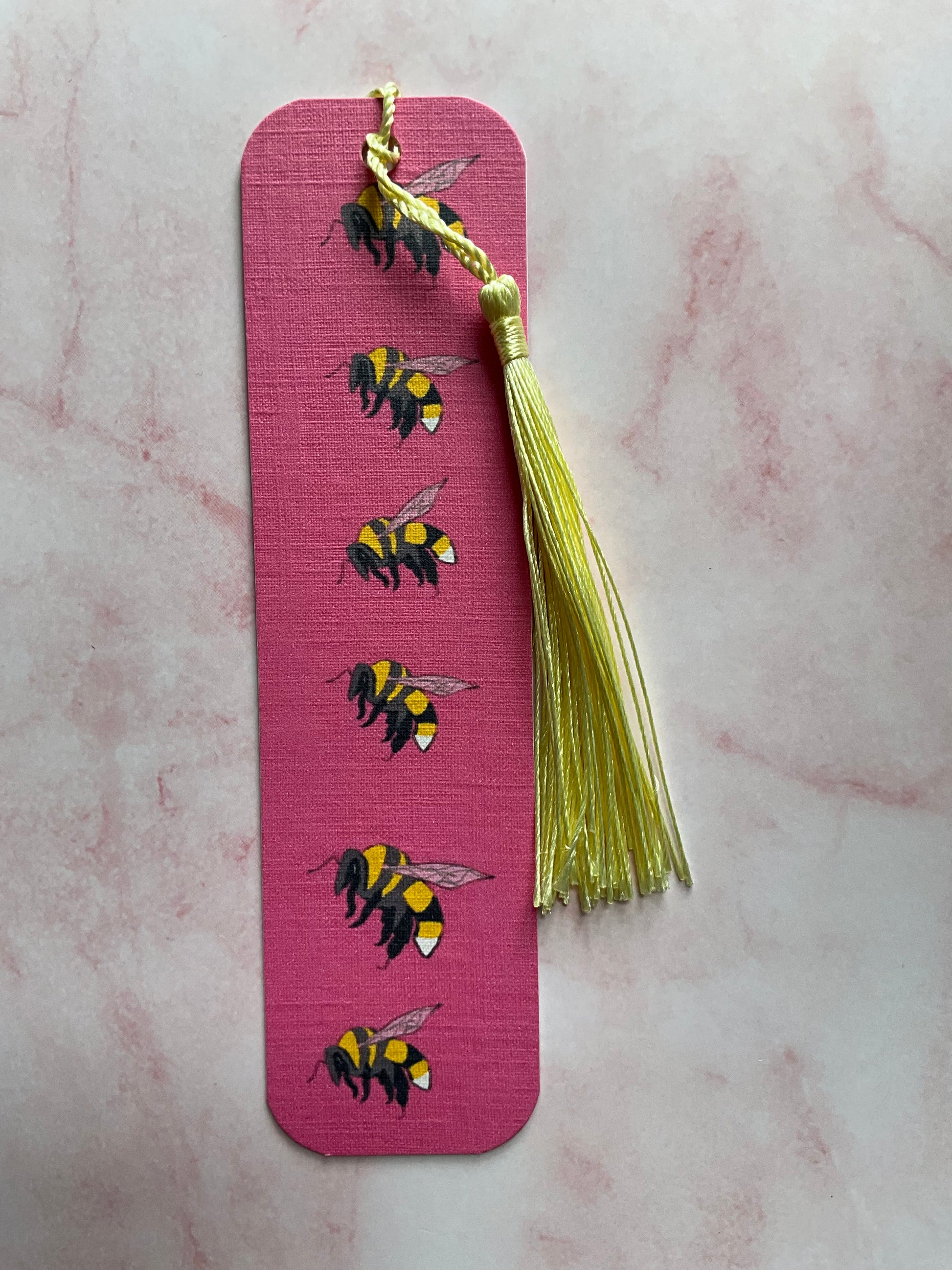 Pink bookmark with bee designs and a gold tassel on a marble surface