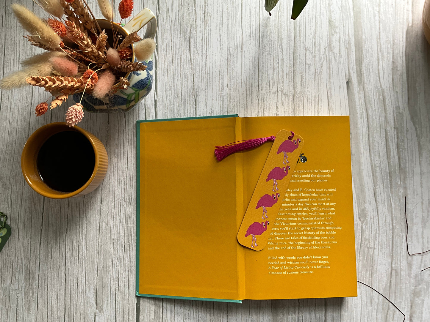Open book with a coourful bookmark on a wooden surface next to a cup of coffee and dried flowers.
