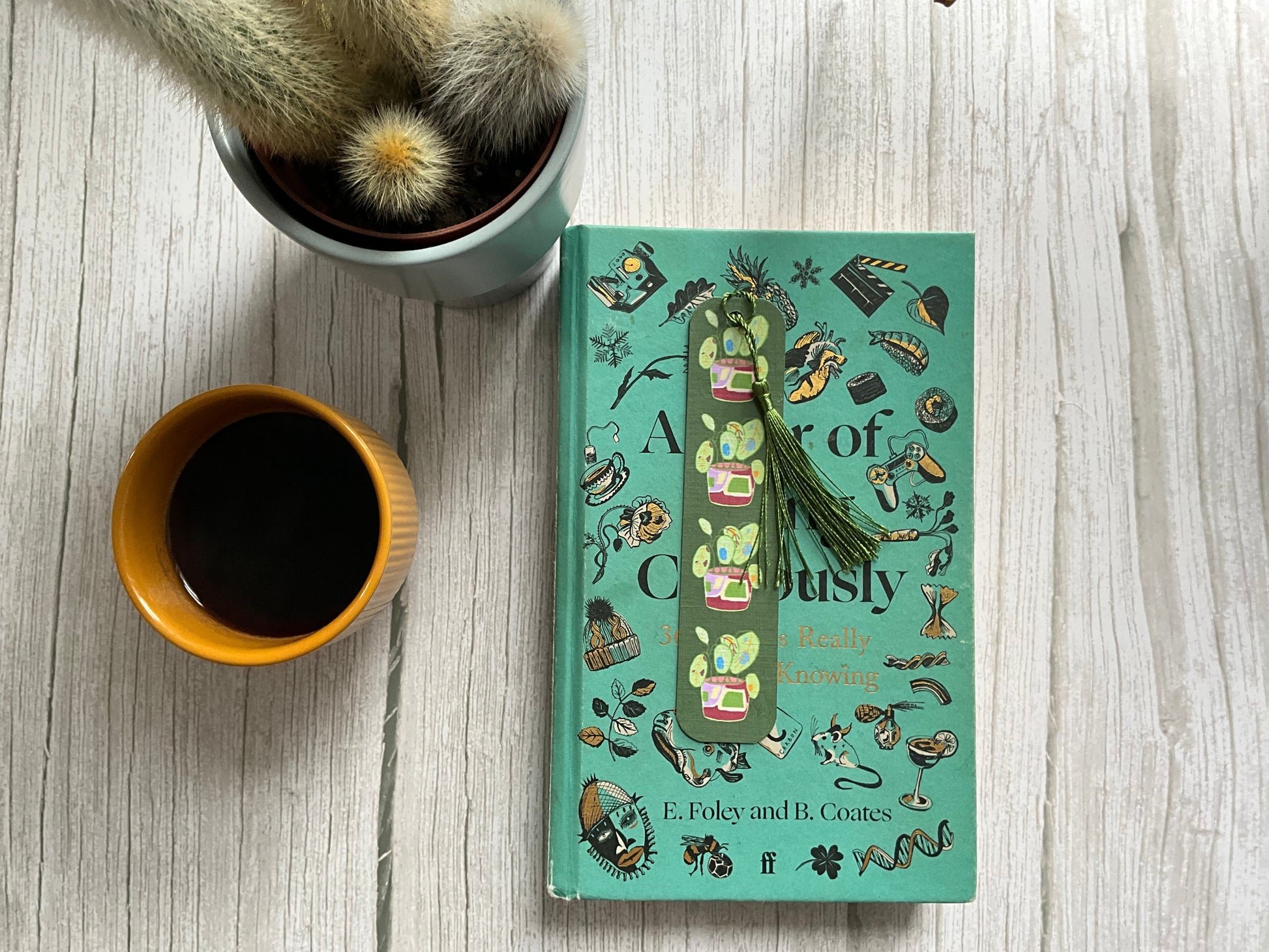 Botanical bookmark featuring leafy plant designs and a green tassel resting on a book, next to a coffee cup and cactus plant