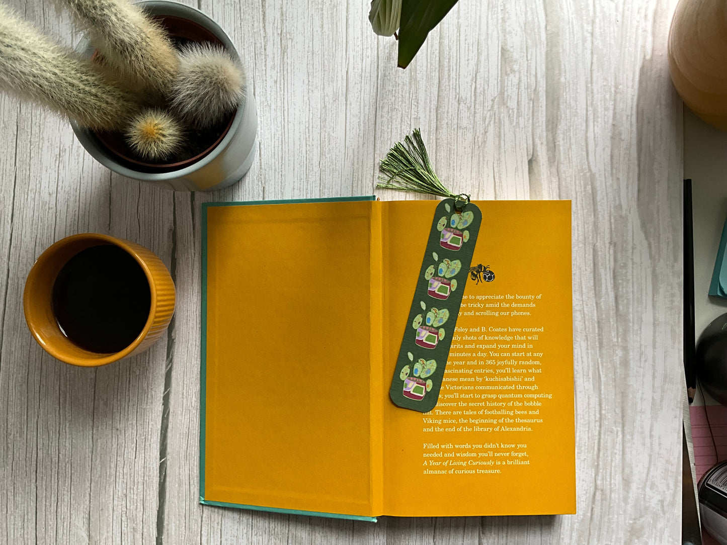 Pot plant design green bookmark on an open book with yellow pages next to a yellow coffee cup and cactus plant