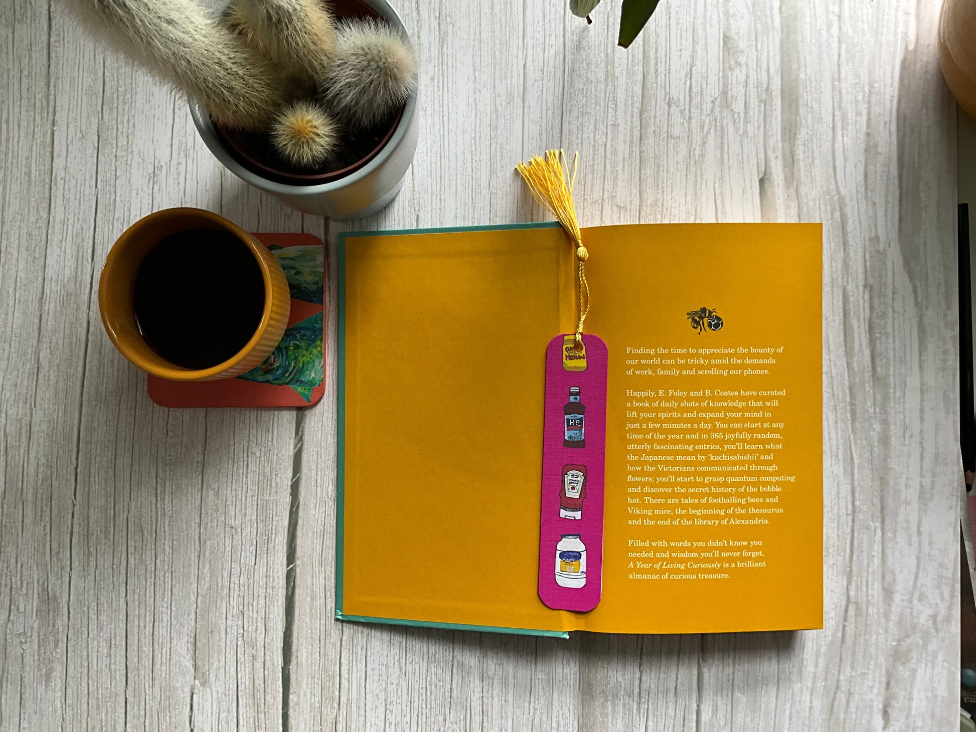 Book with a pink colourful bookmark on a wooden surface next to a cup of coffee and a cactus plant.