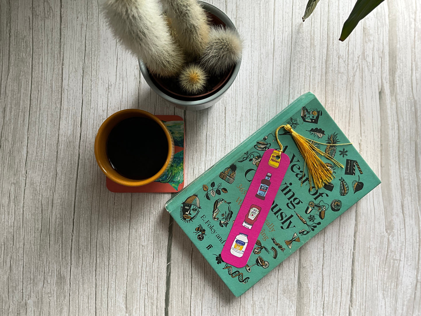 A pink bookmark with a sauce print design resting on a book next to a coffee cup and a cactus plant