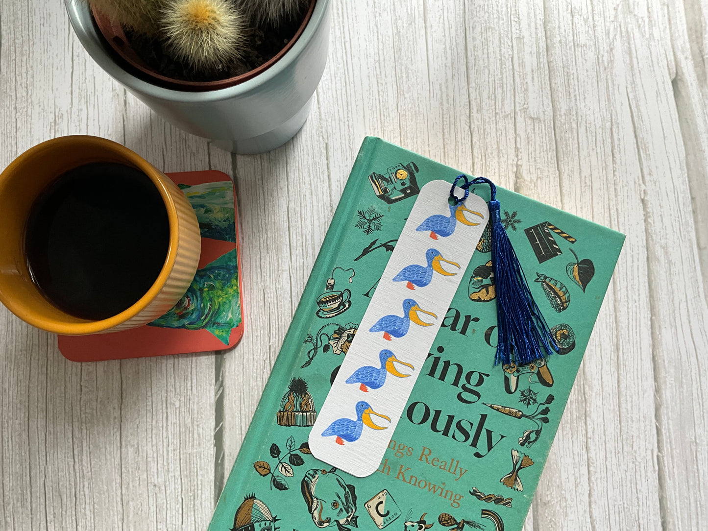 White bookmark with blue pelican print and a blue tassel, resting on a book with a coffee cup and cactus plant next to it. 