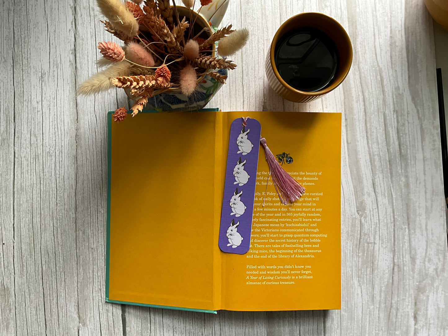 Open book with a purple bookmark featuring rabbit illustrations on a wooden surface with a cup of coffee and dried plants.