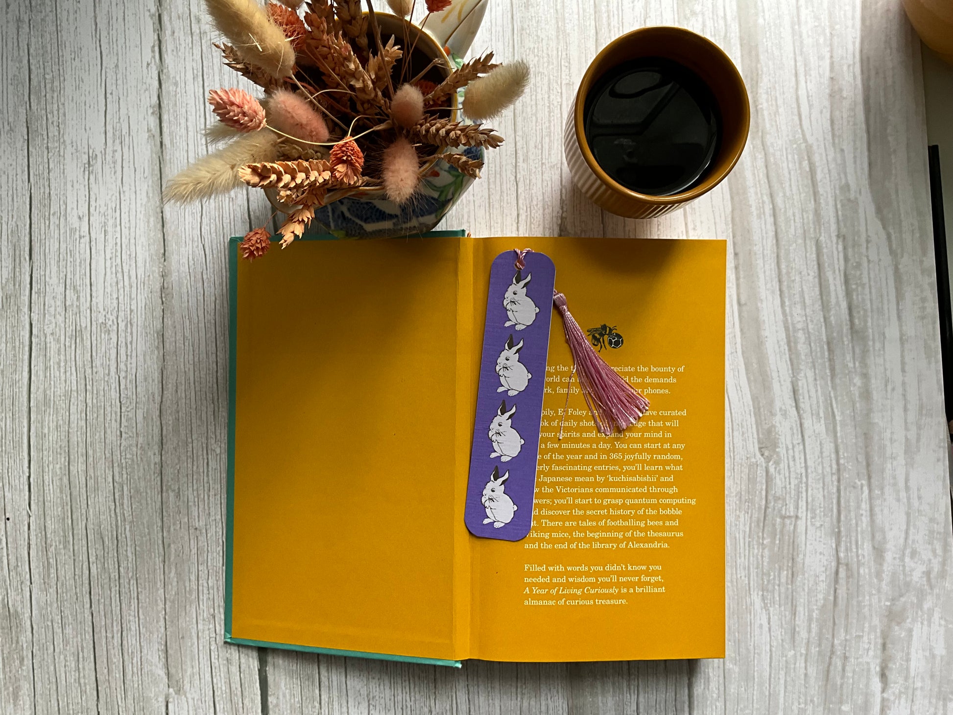Open book with a purple bookmark featuring rabbit illustrations on a wooden surface with a cup of coffee and dried plants.