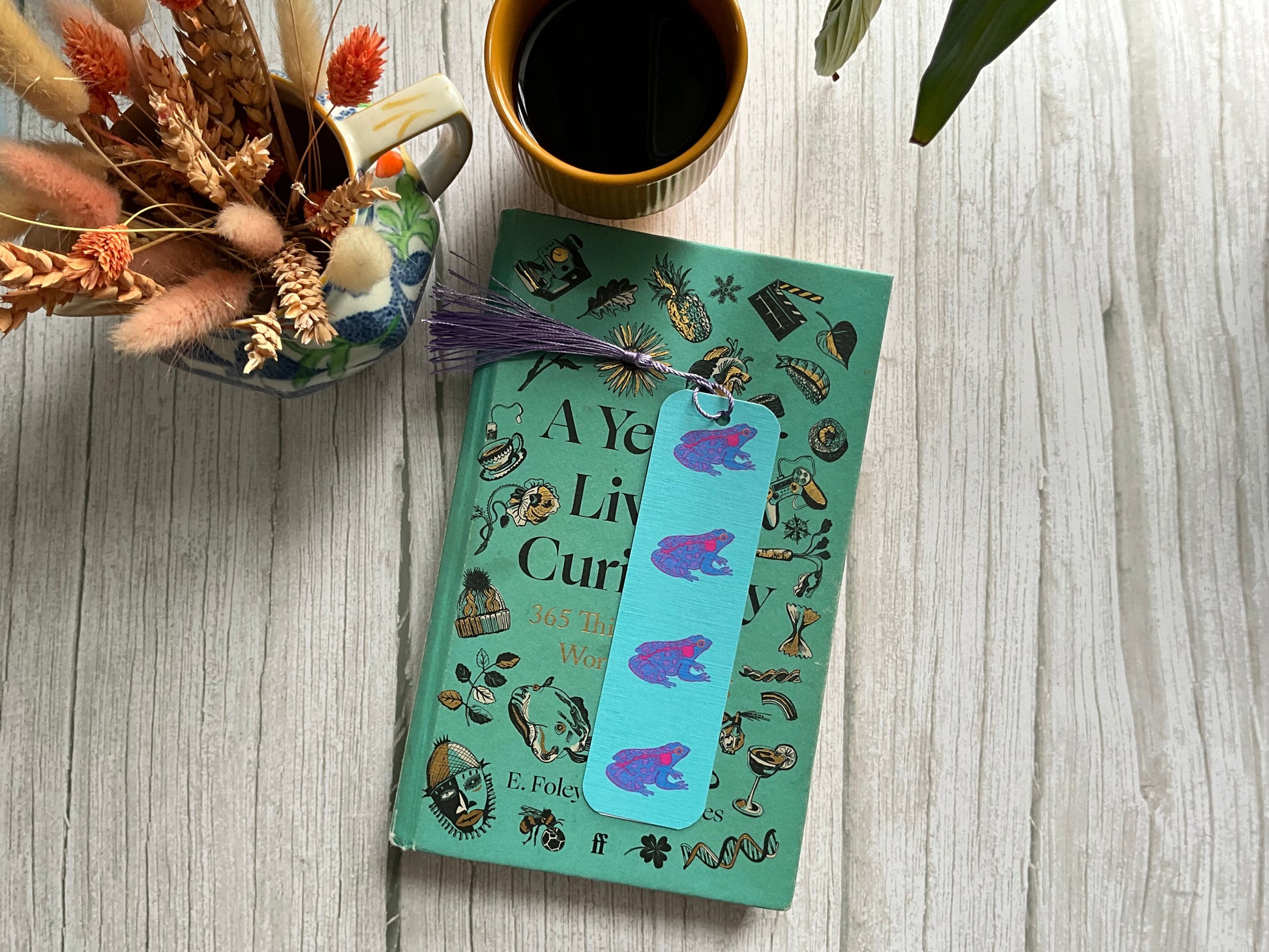Blue frog patterned bookmark with tassel resting on a teal book on a table with dried flowers and a cup of coffee