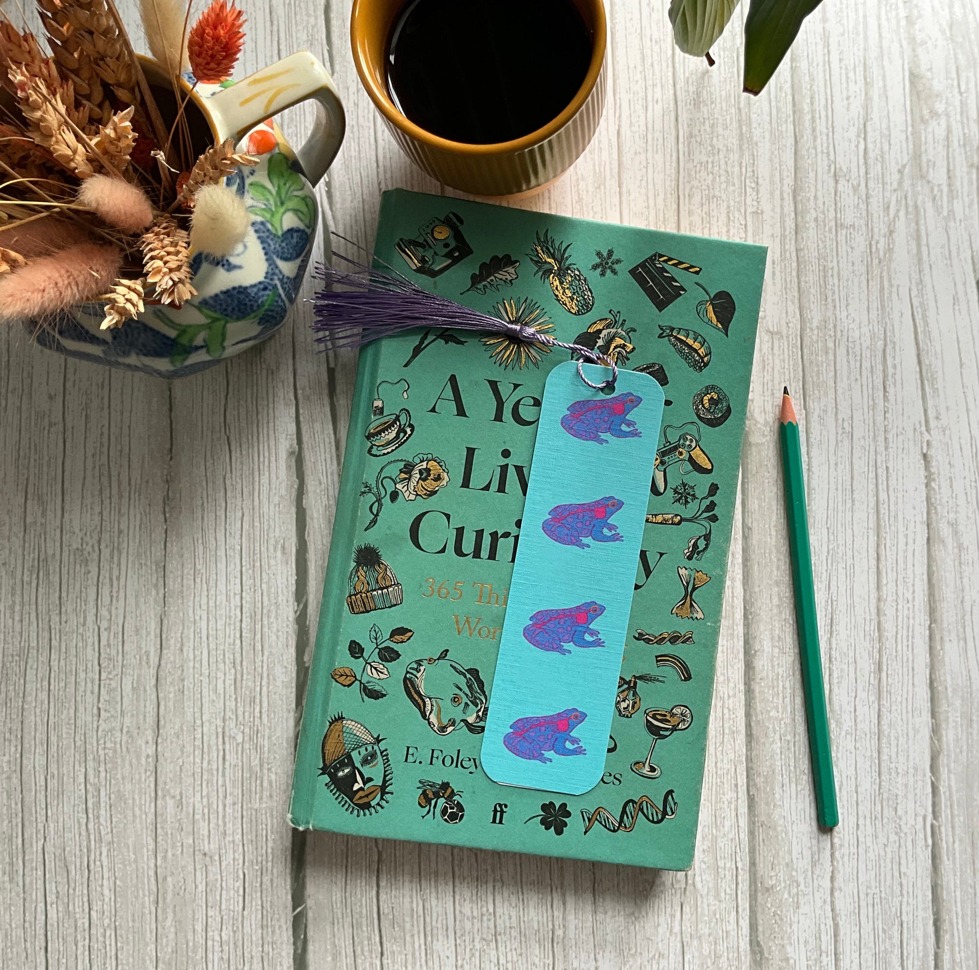 Book with a blue bookmark with a purple frog design resting on a wooden table with a vase of dried flowers and yellow coffee cup